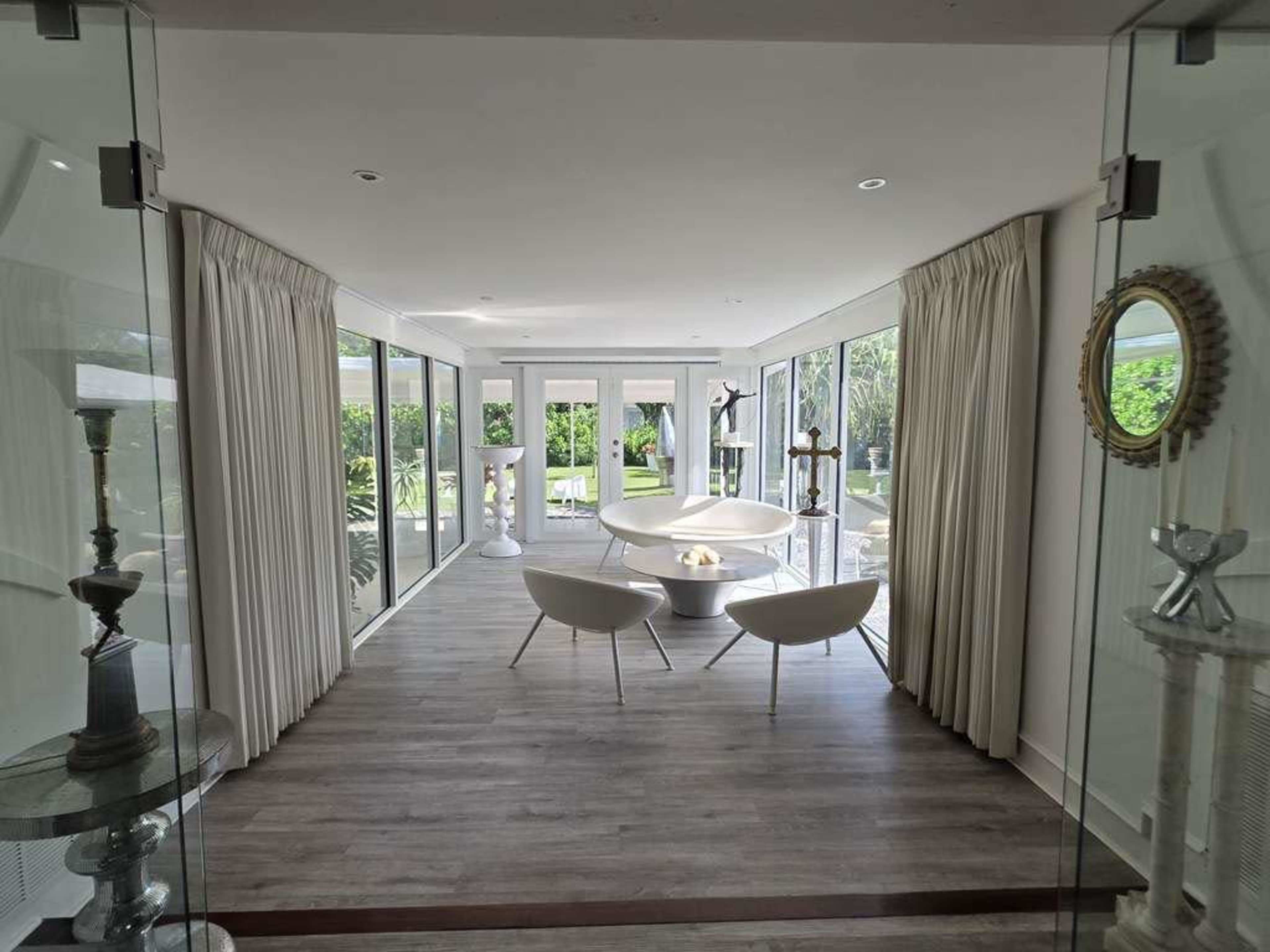 The image shows a modern, open-concept living space with large glass windows, white furniture, and light-colored curtains, leading to an outdoor garden.
