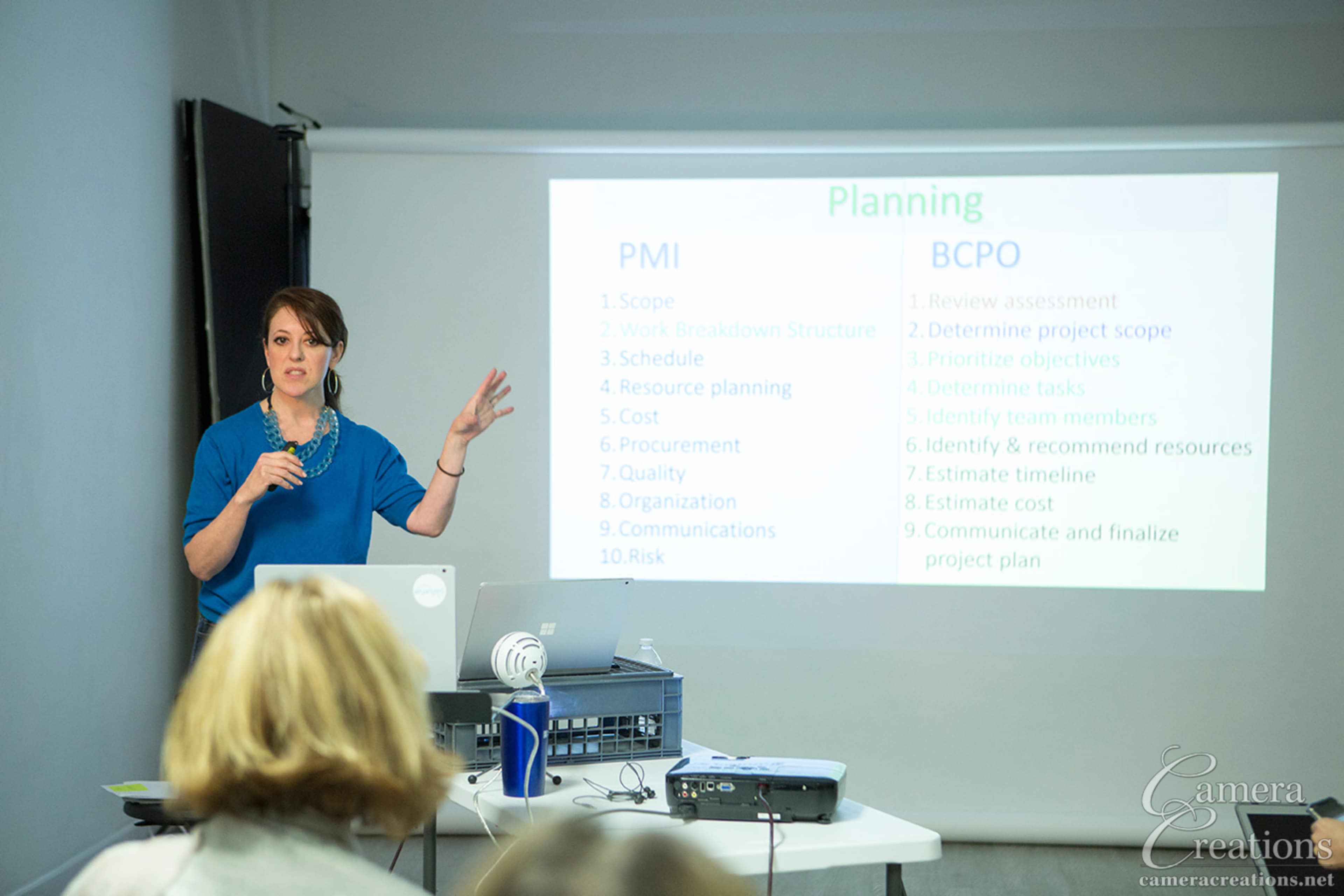 A presenter stands in front of an audience, gesturing towards a projector screen displaying a list of planning steps for project management.