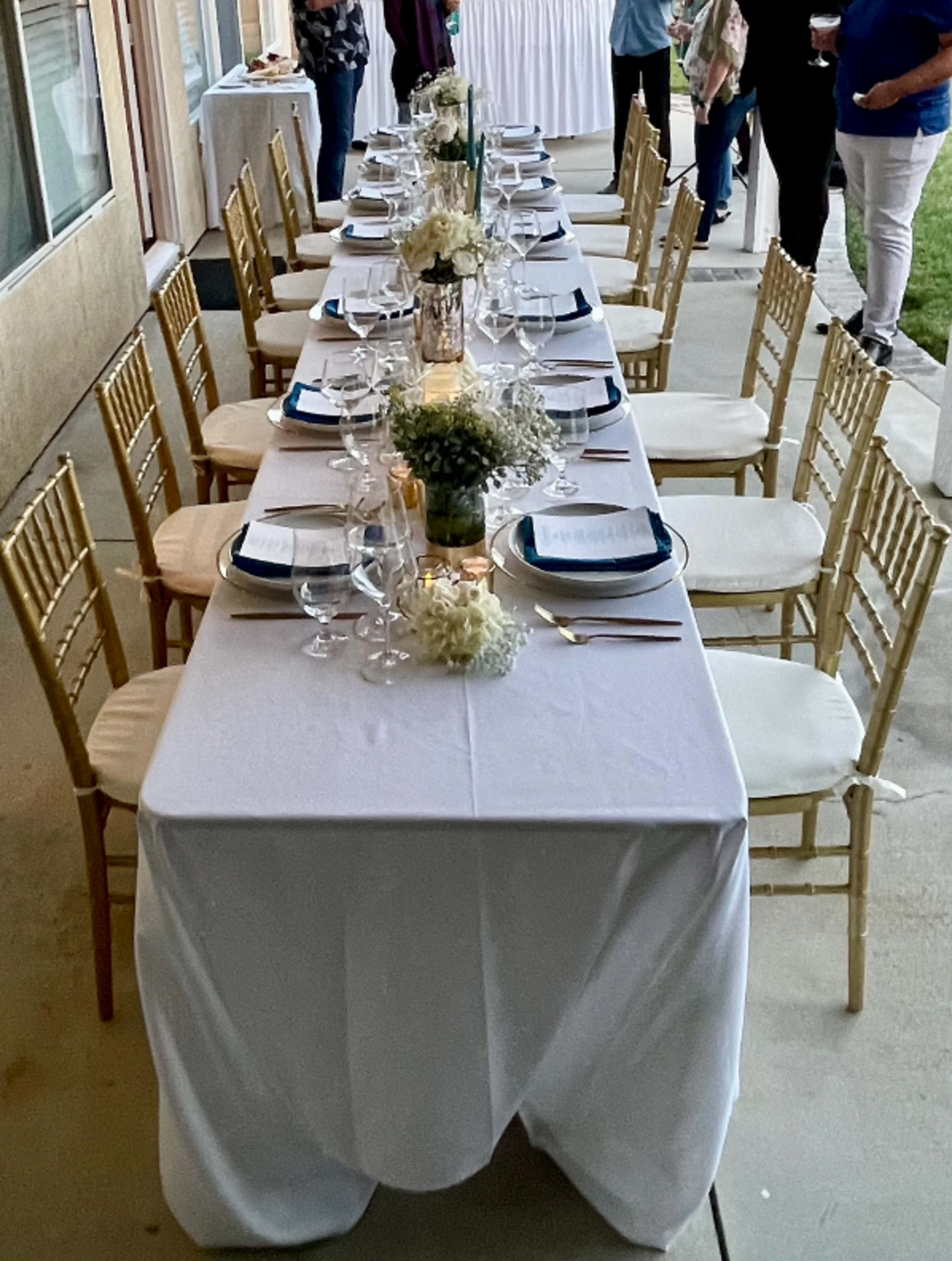 A long, elegantly set dining table with white tablecloths, plates, glasses, and floral centerpieces is arranged outdoors, while guests are mingling in the background.