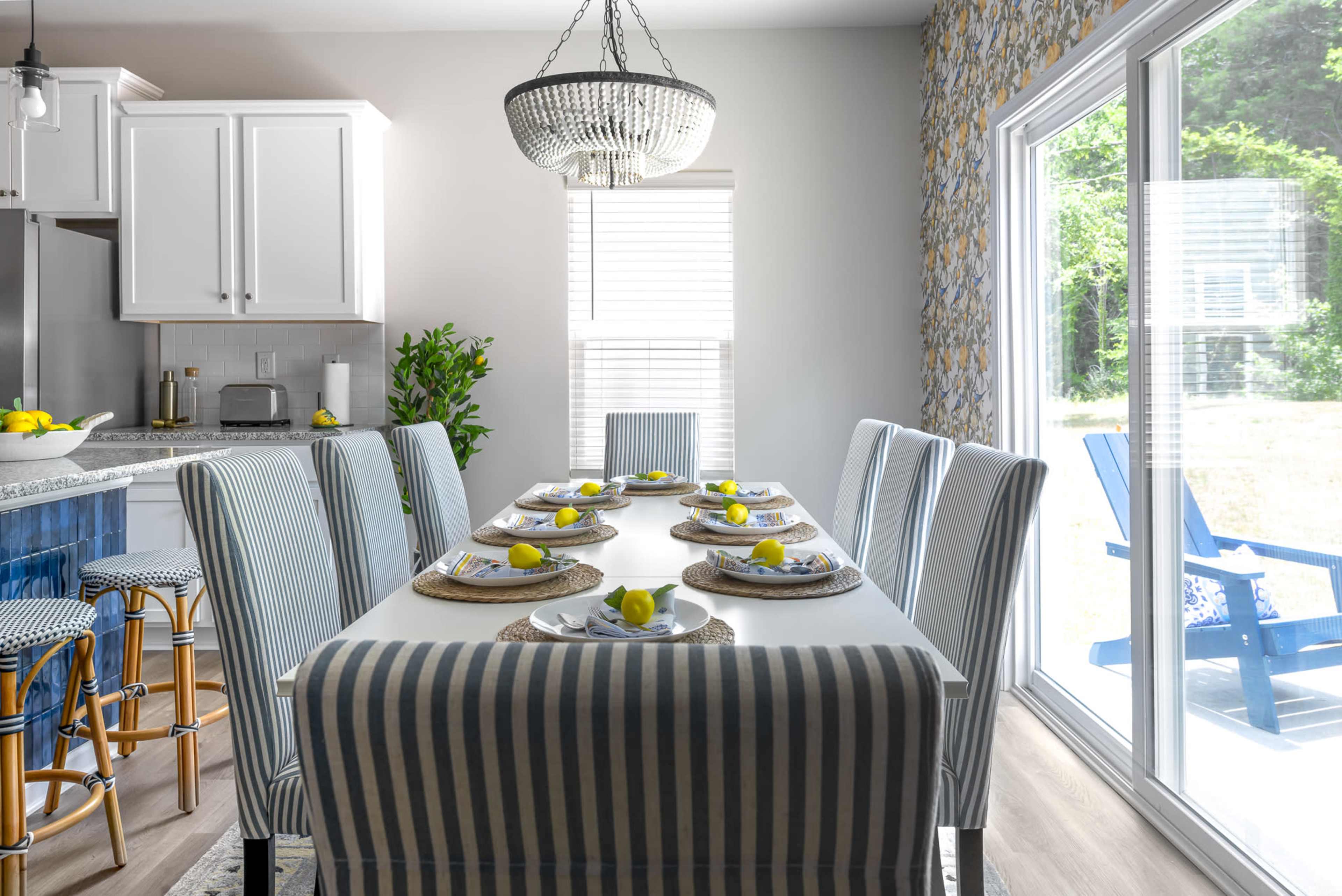 A dining table set for eight with striped chairs, decorative plates, and lemons, is positioned near a large window that leads to an outdoor space.