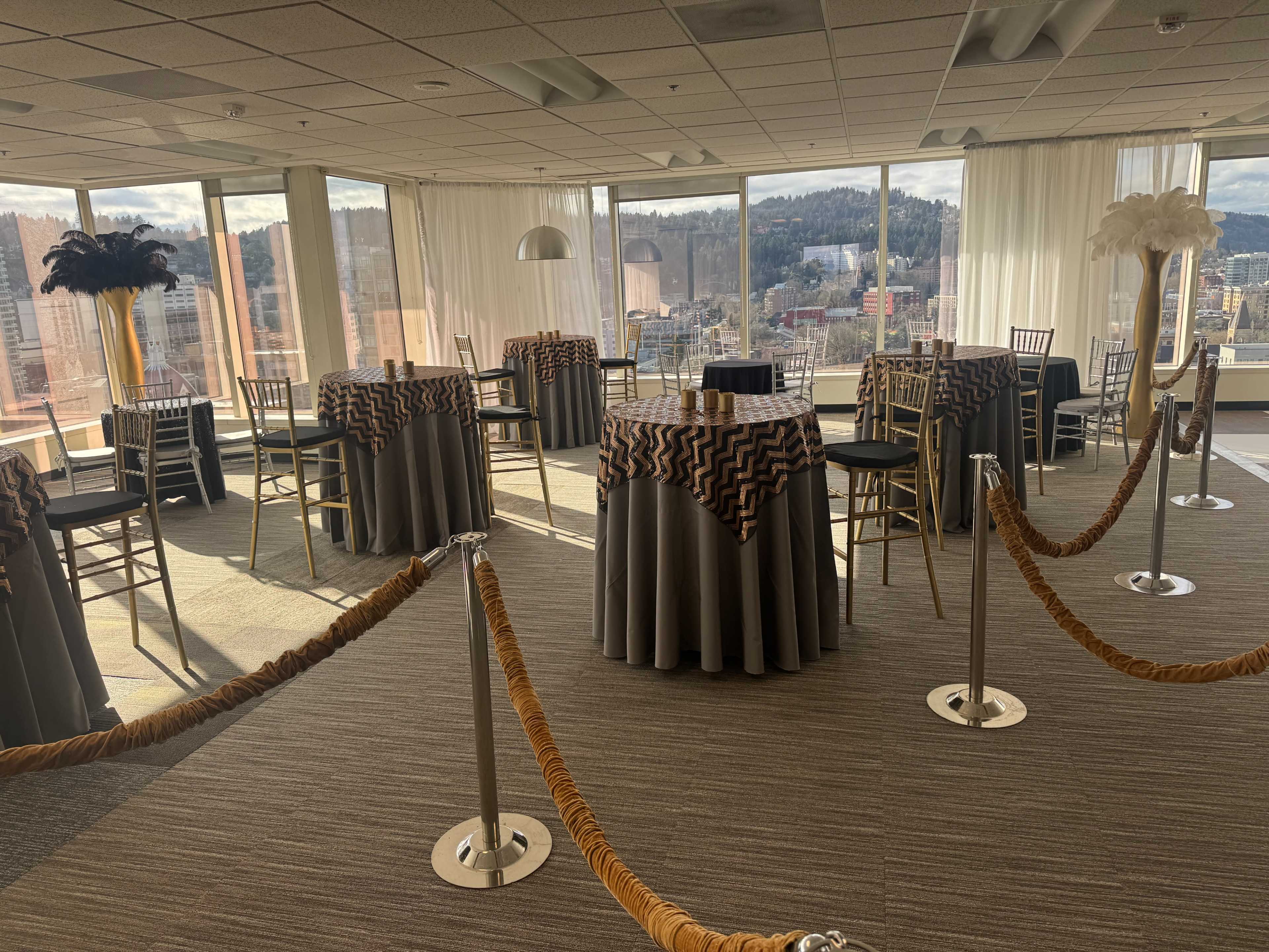 The image shows a spacious event room with tall windows overlooking a cityscape, featuring several round tables covered with decorative tablecloths and surrounded by gold rope barriers.