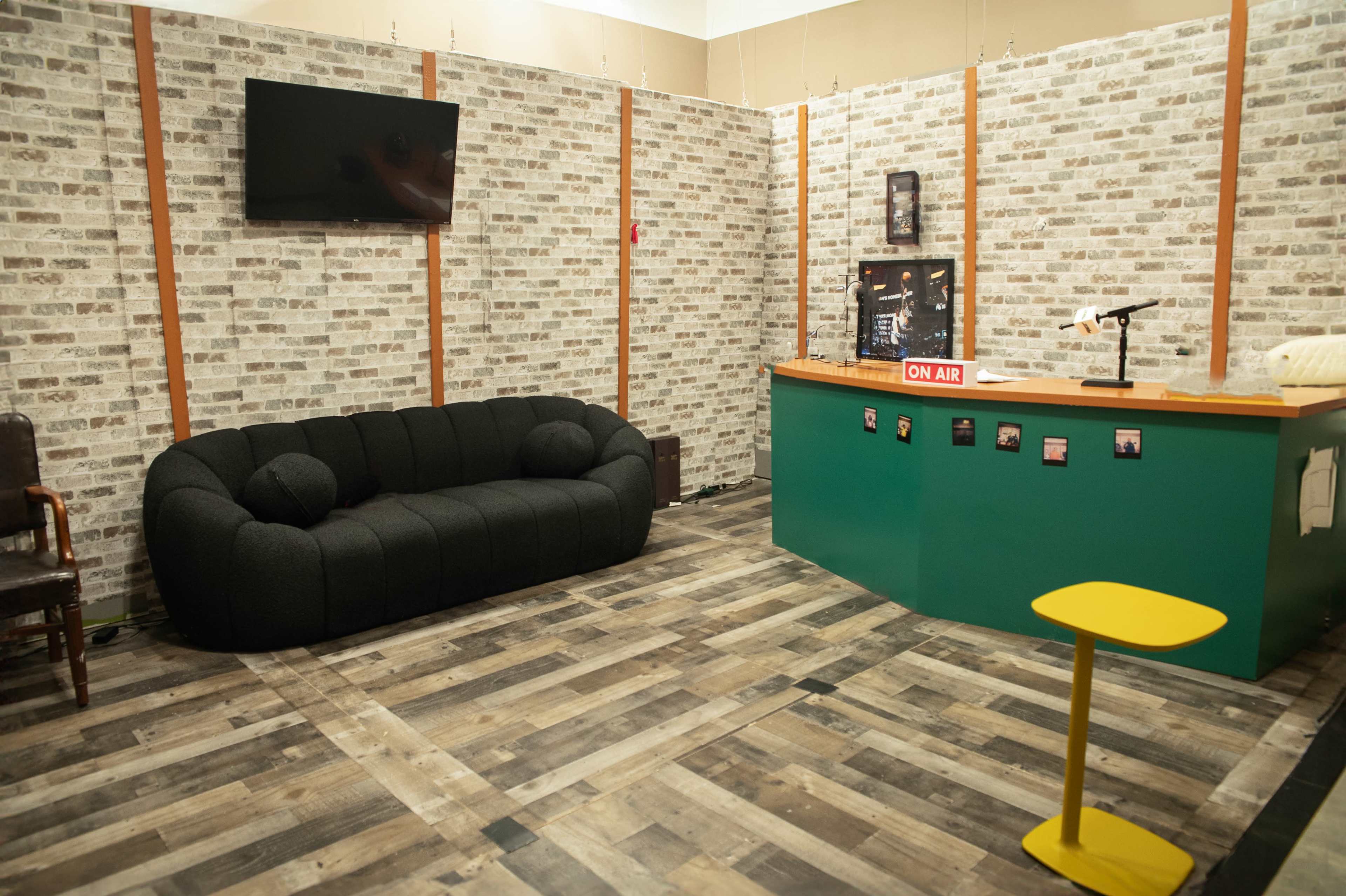 The image shows a studio-like space featuring a black sofa, a green reception counter with an "ON AIR" sign, and a wall decorated with brick-patterned wallpaper.