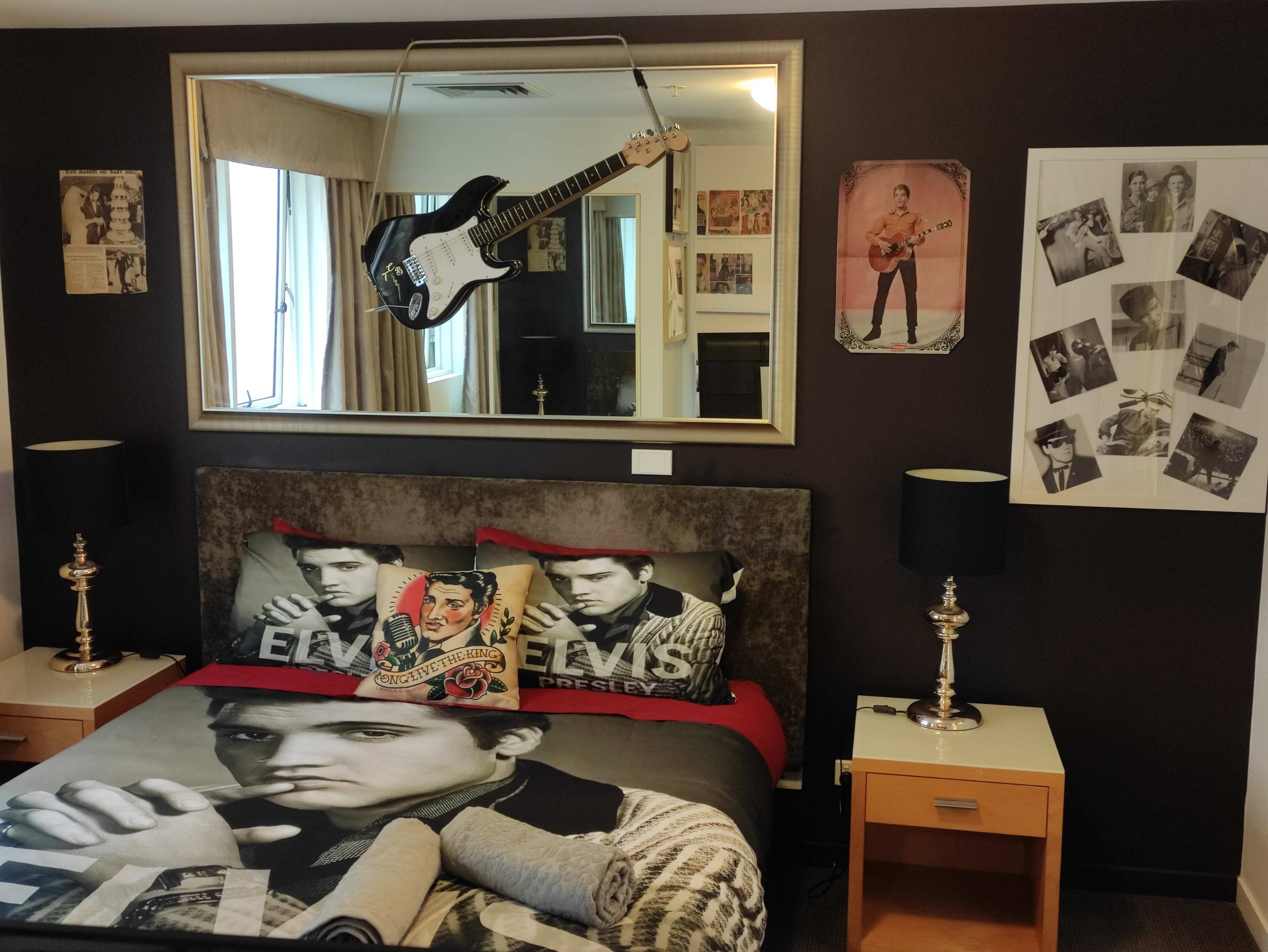 A bedroom featuring an Elvis Presley-themed bedspread, framed pictures of Elvis on the walls, and a guitar suspended above a mirrored headboard.
