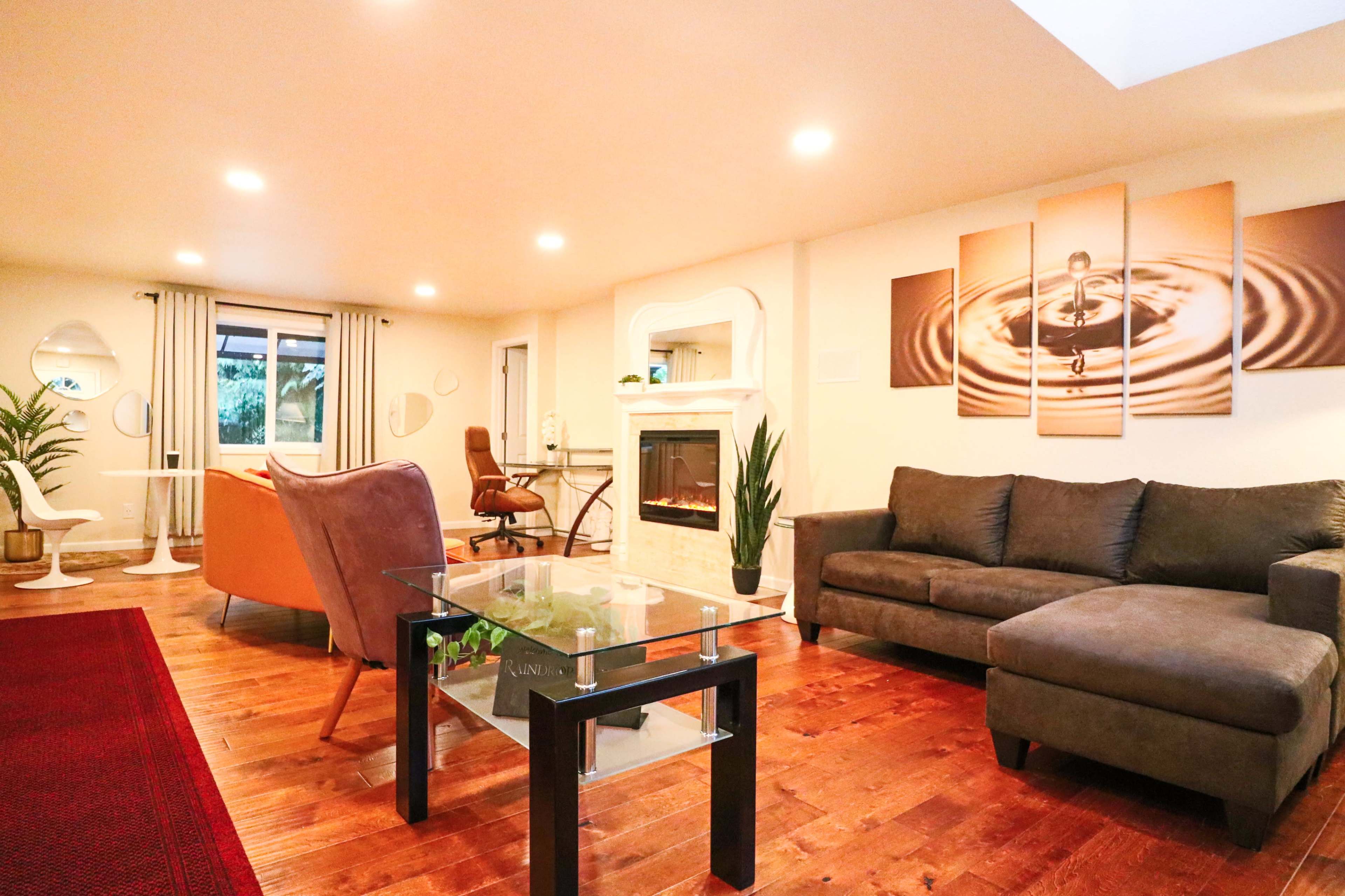 The image shows a modern living room featuring a gray couch, a glass coffee table, a fireplace, and an arrangement of chairs around a central area.