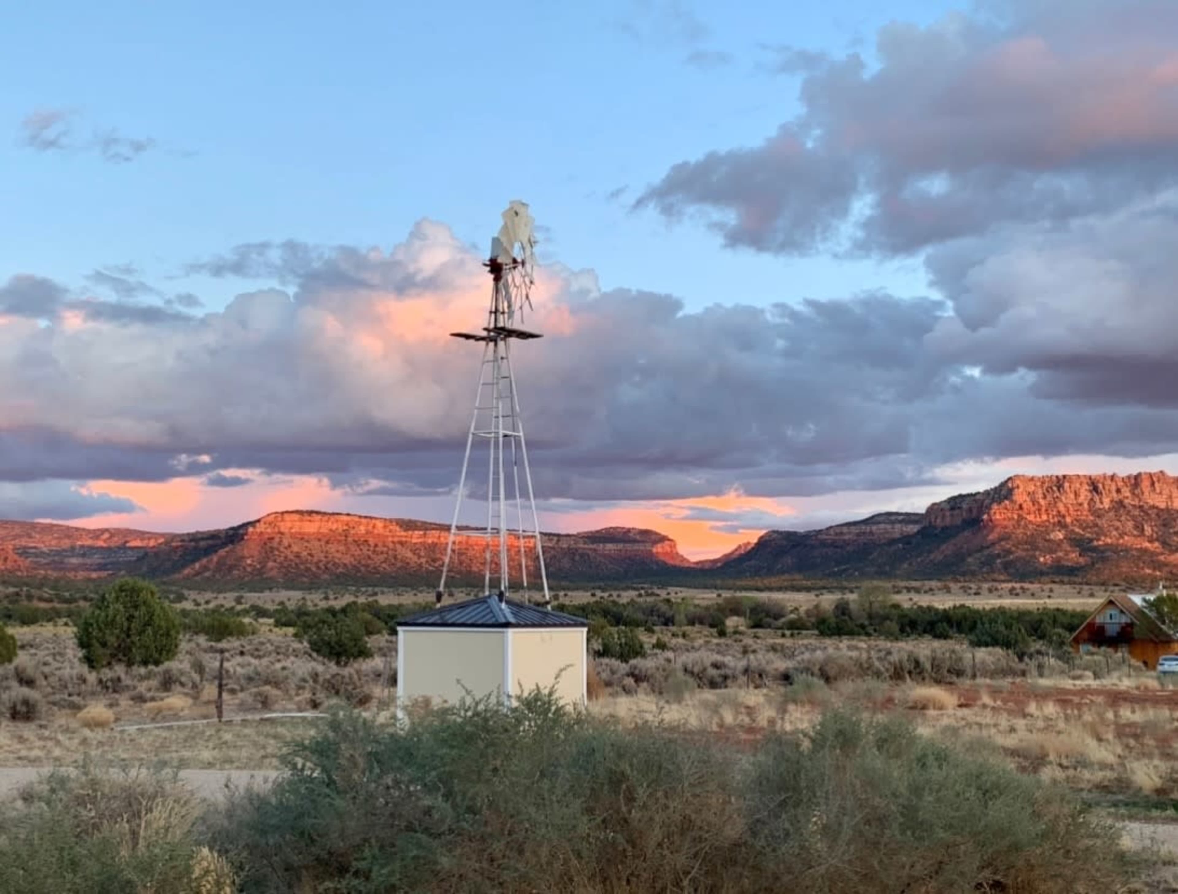 A windmill stands on a flat landscape with distant mountains under a colorful sky filled with clouds at sunset.