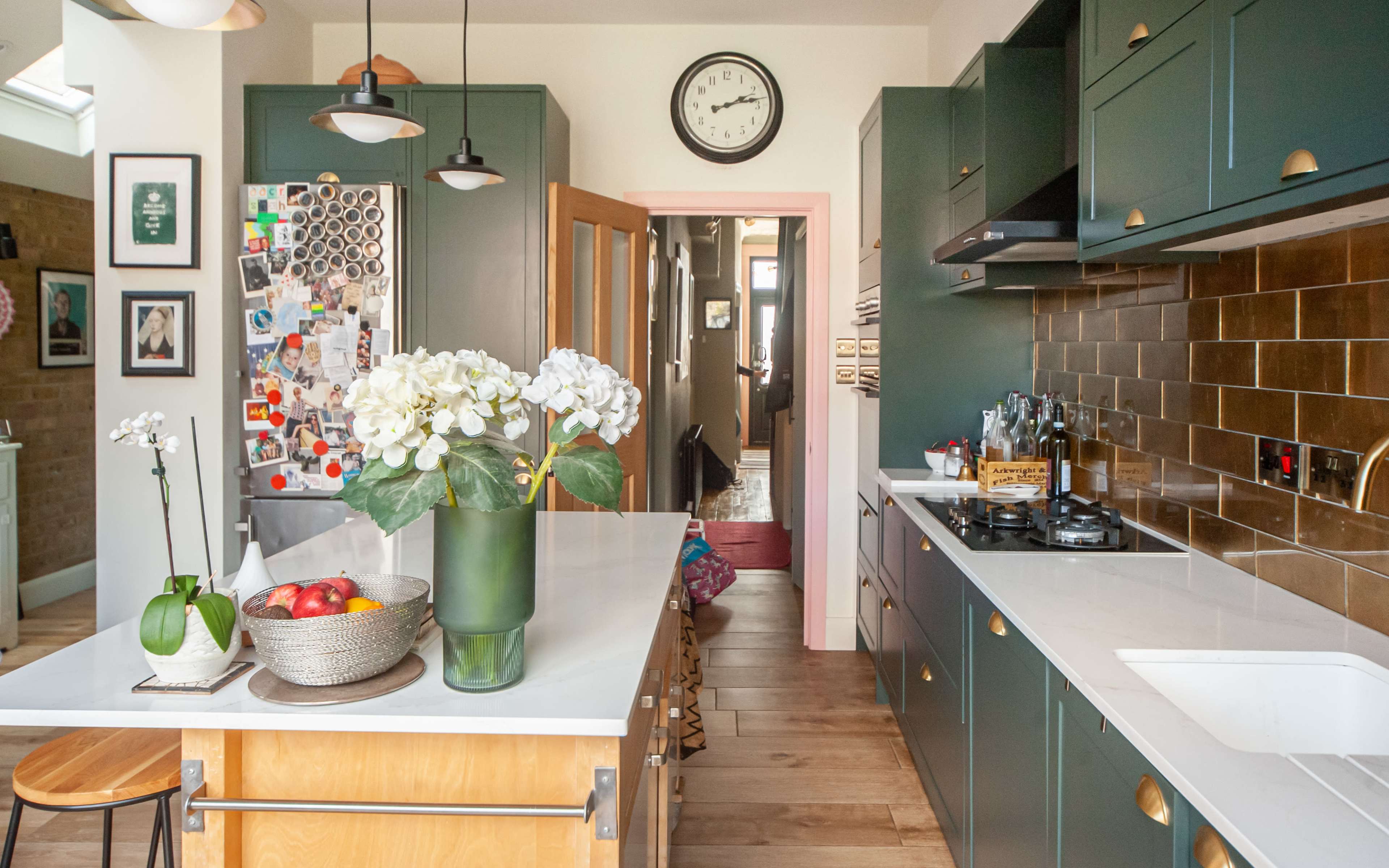 The kitchen features dark green cabinetry, a white countertop, and a clock on the wall, with a fruit bowl and a vase of flowers on the island.