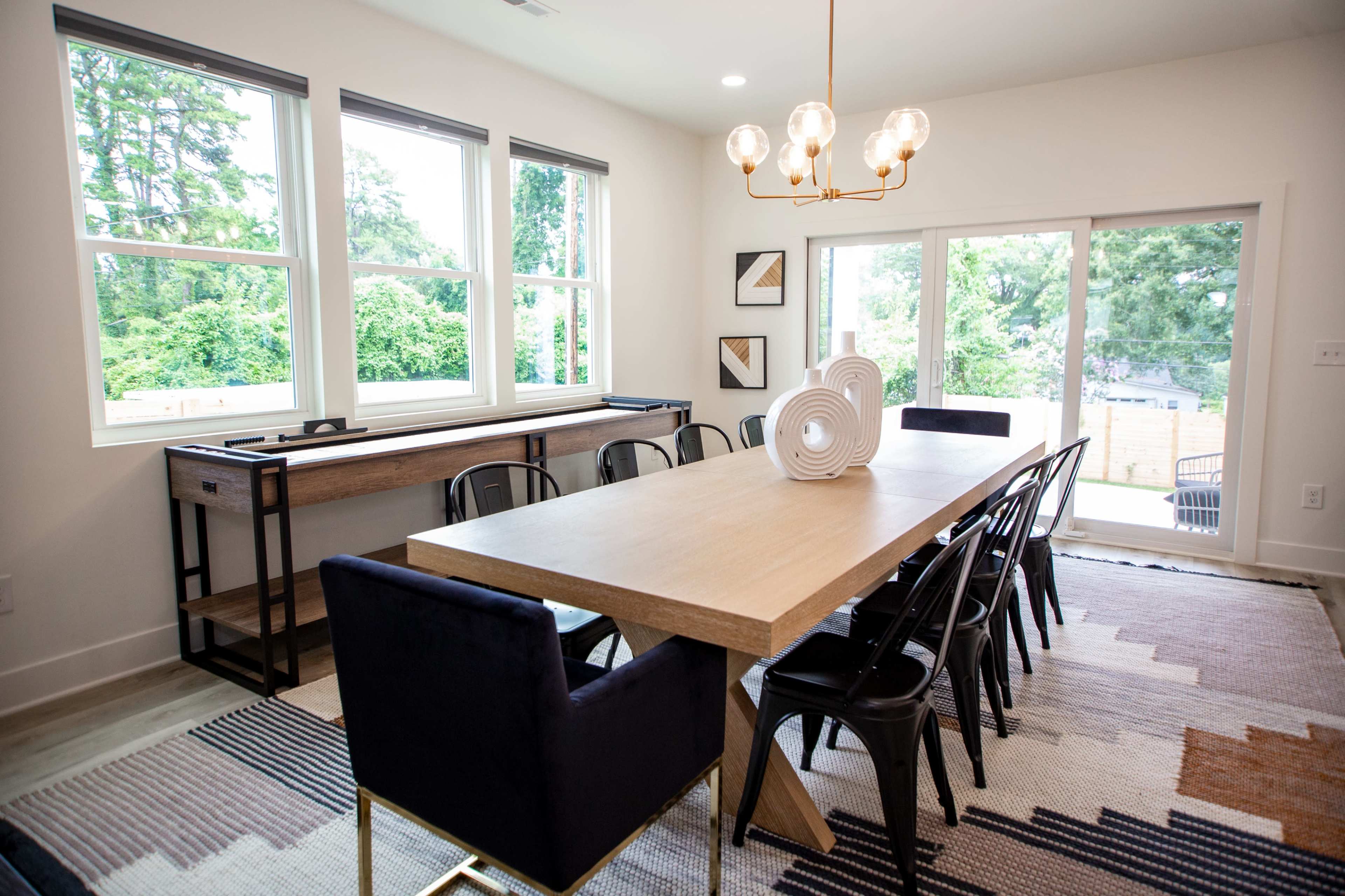 A dining room features a large wooden table with black chairs, a patterned rug, and windows overlooking greenery.