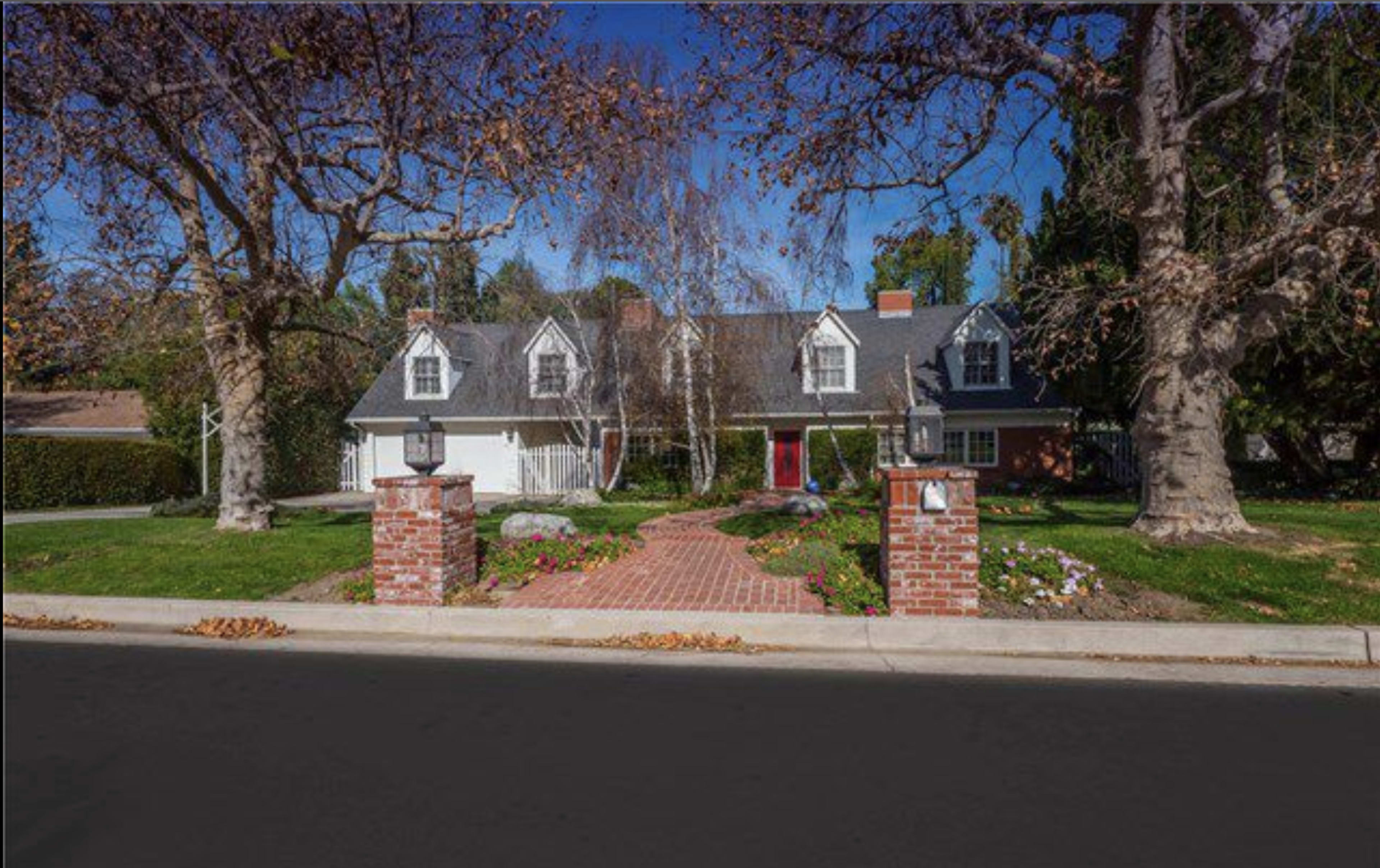 A two-story house with a sloped roof, multiple dormer windows, and a red front door is set behind a brick walkway and landscaped front yard.