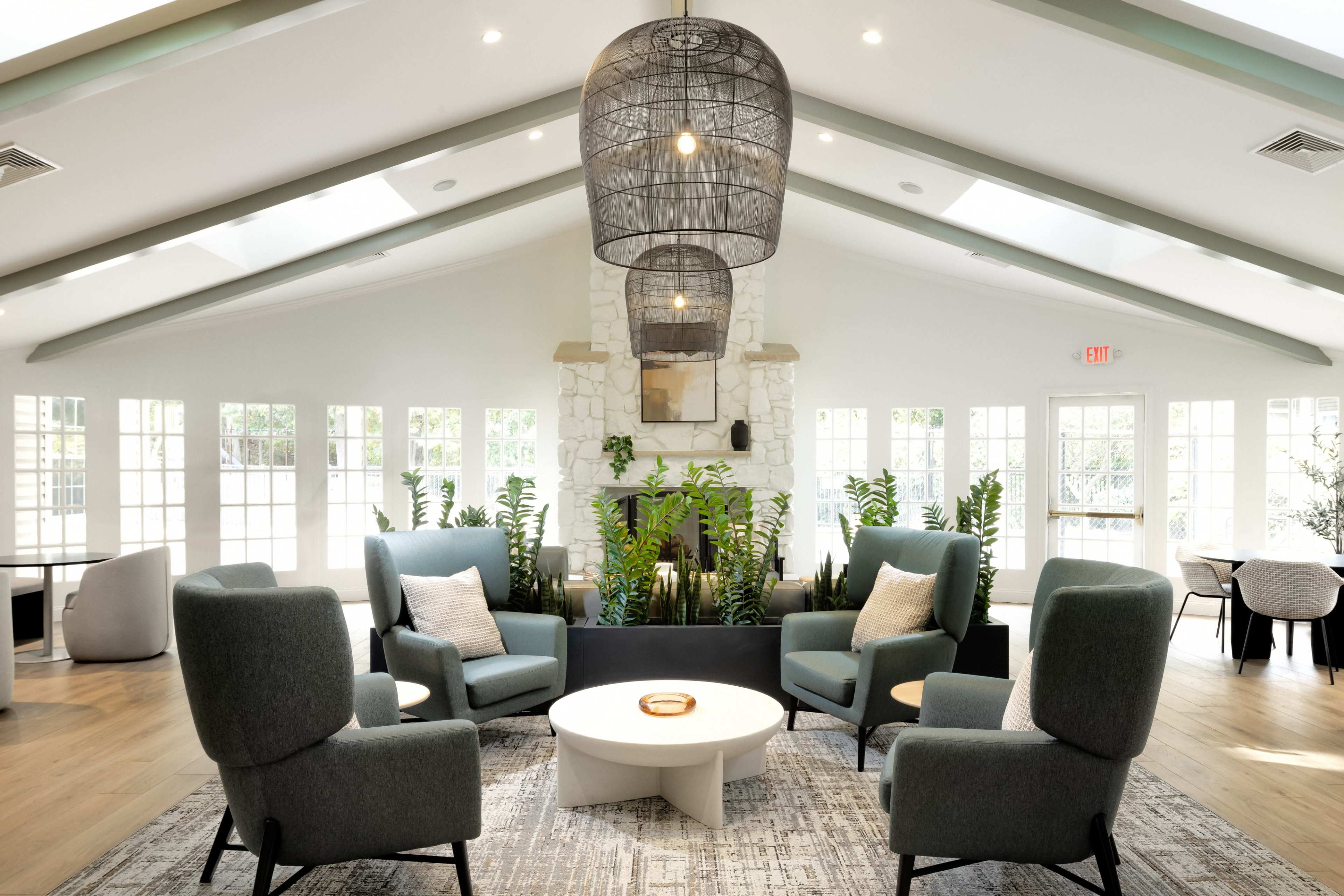 The image shows a modern indoor lounge area featuring four armchairs arranged around a circular coffee table, with large windows allowing natural light and greenery visible in the background.