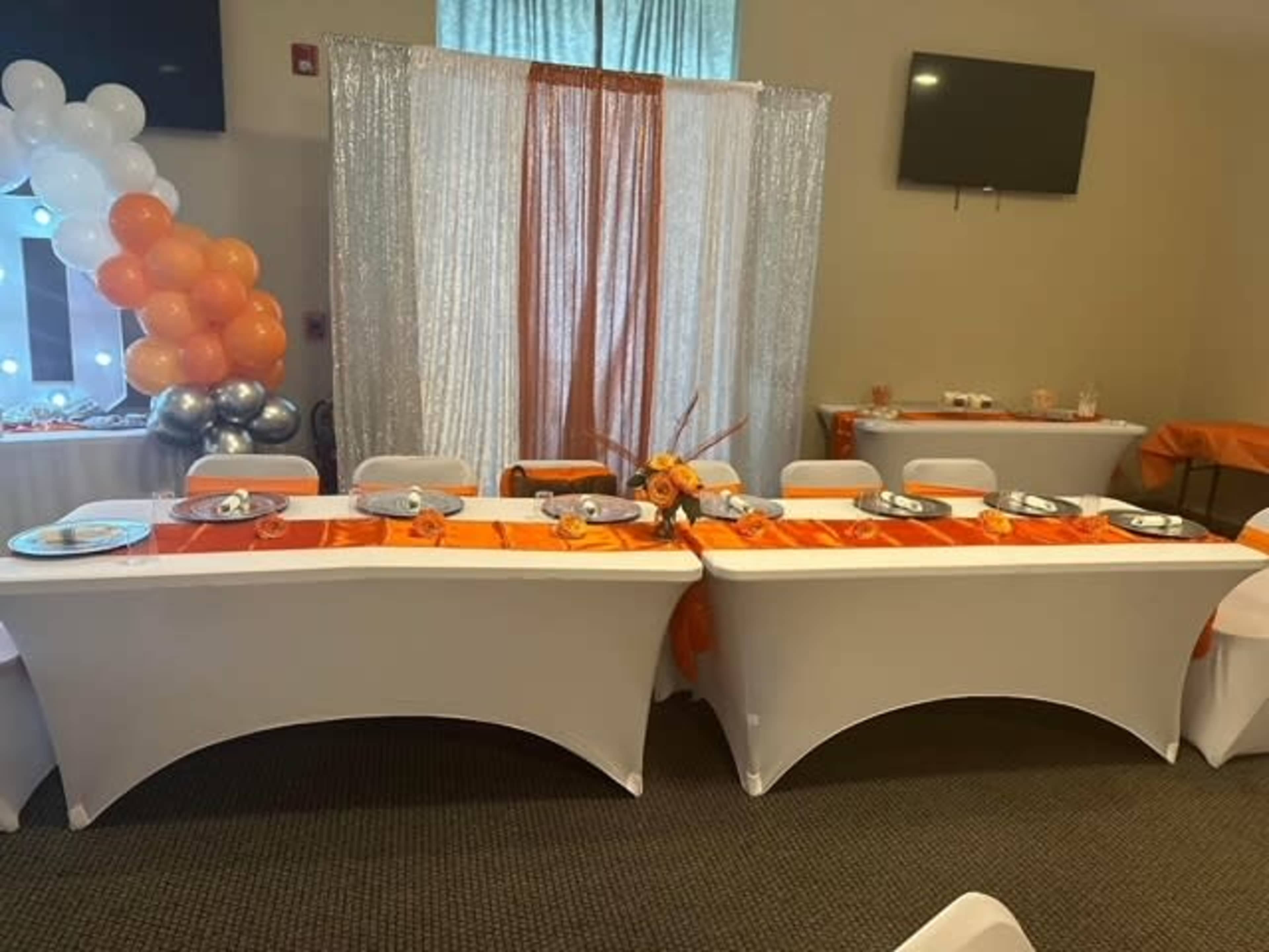 A decorated event space featuring two tables draped in white cloth, adorned with orange table runners, and accented with floral arrangements and balloons.