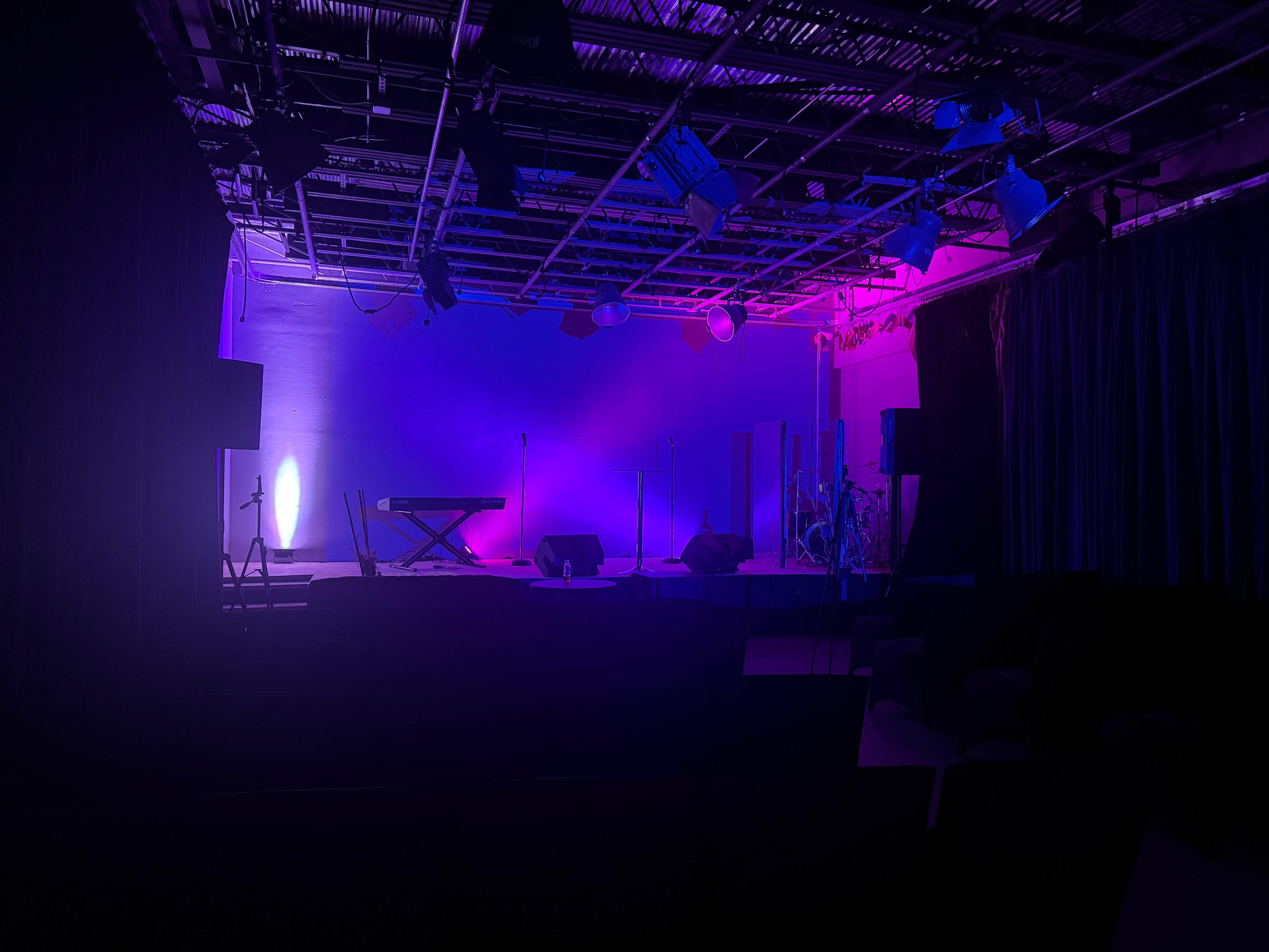 A dimly lit stage with a keyboard, microphones, and drum kit, illuminated by purple and blue lights.