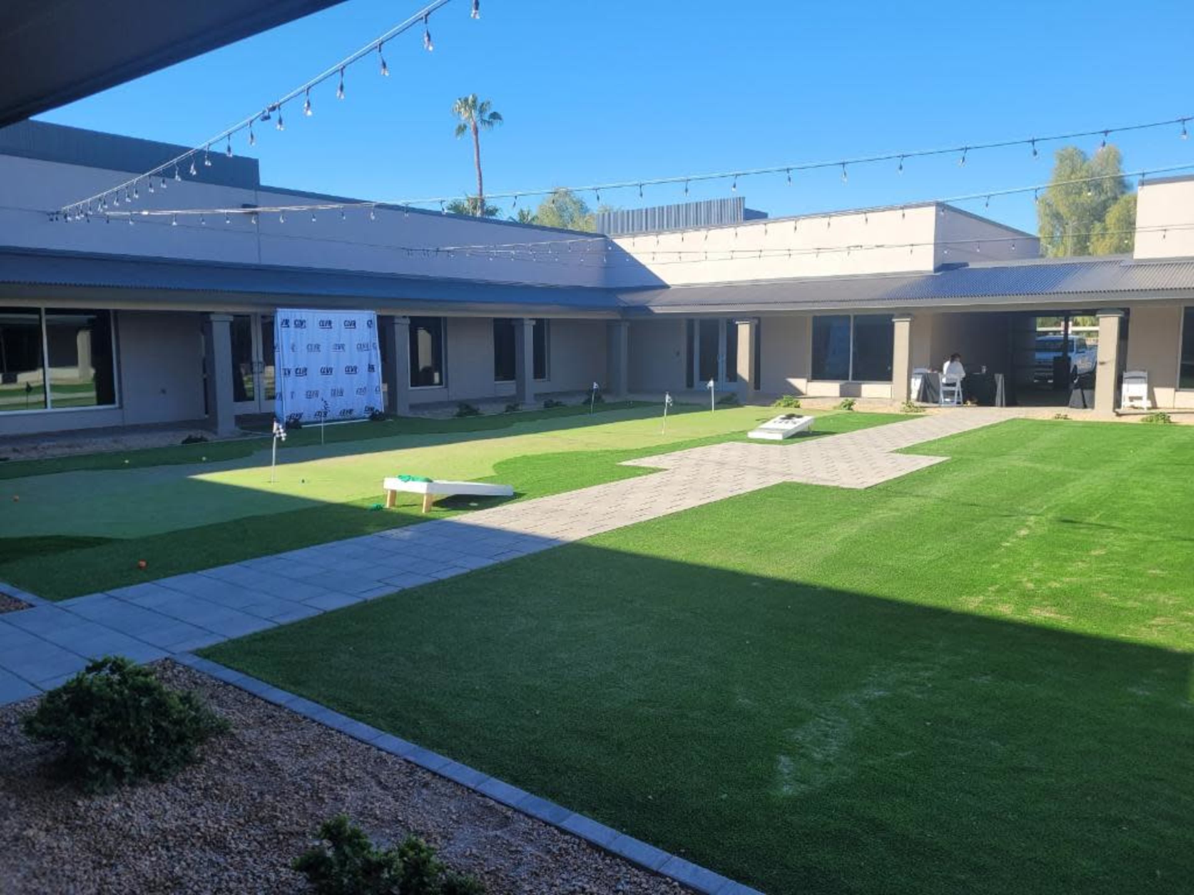 A landscaped courtyard featuring a green lawn, a paved pathway, and a backdrop with a logo, all surrounded by buildings.
