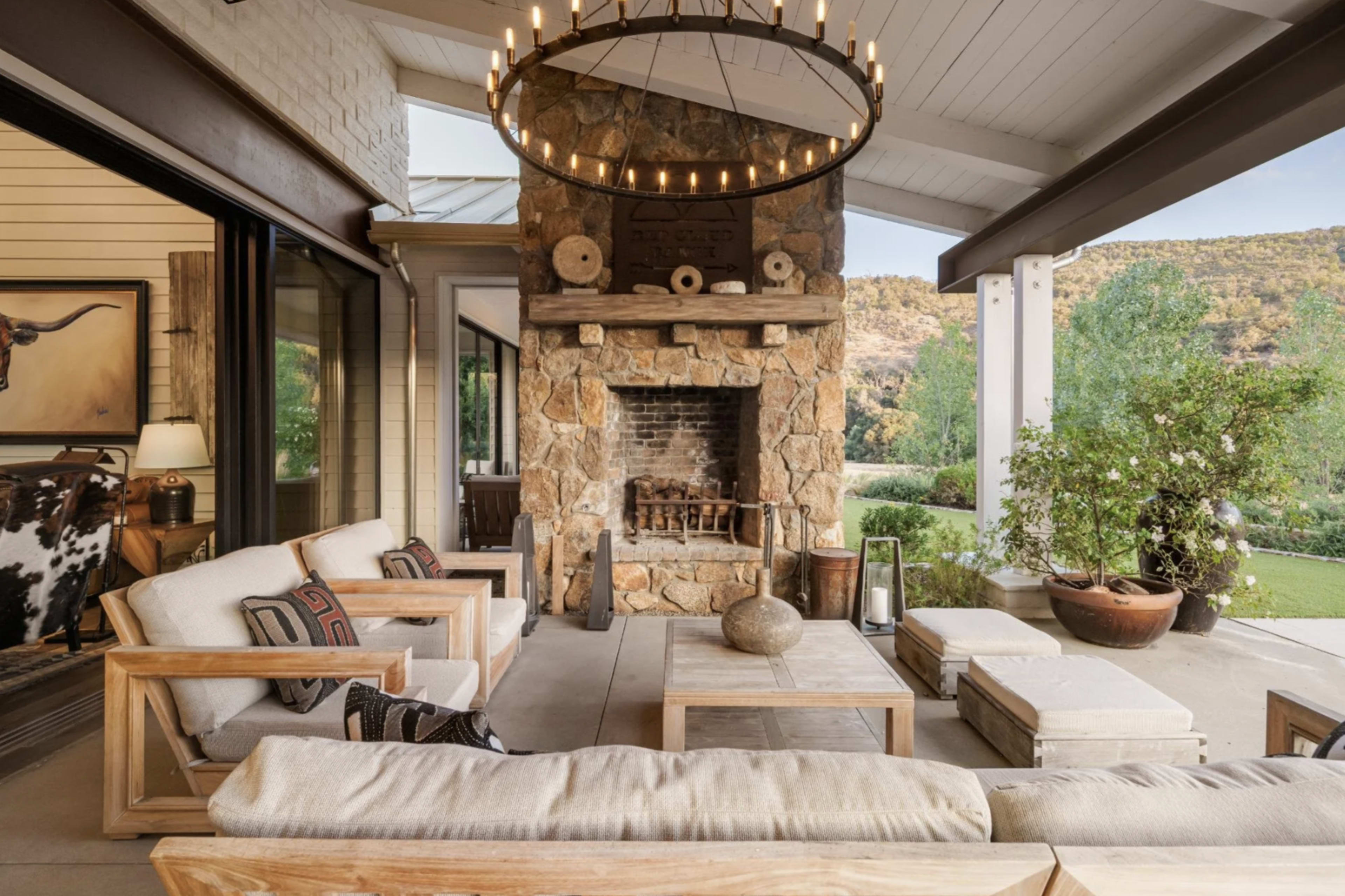 The image shows a spacious outdoor living area with a stone fireplace, wooden furniture, and a large chandelier.