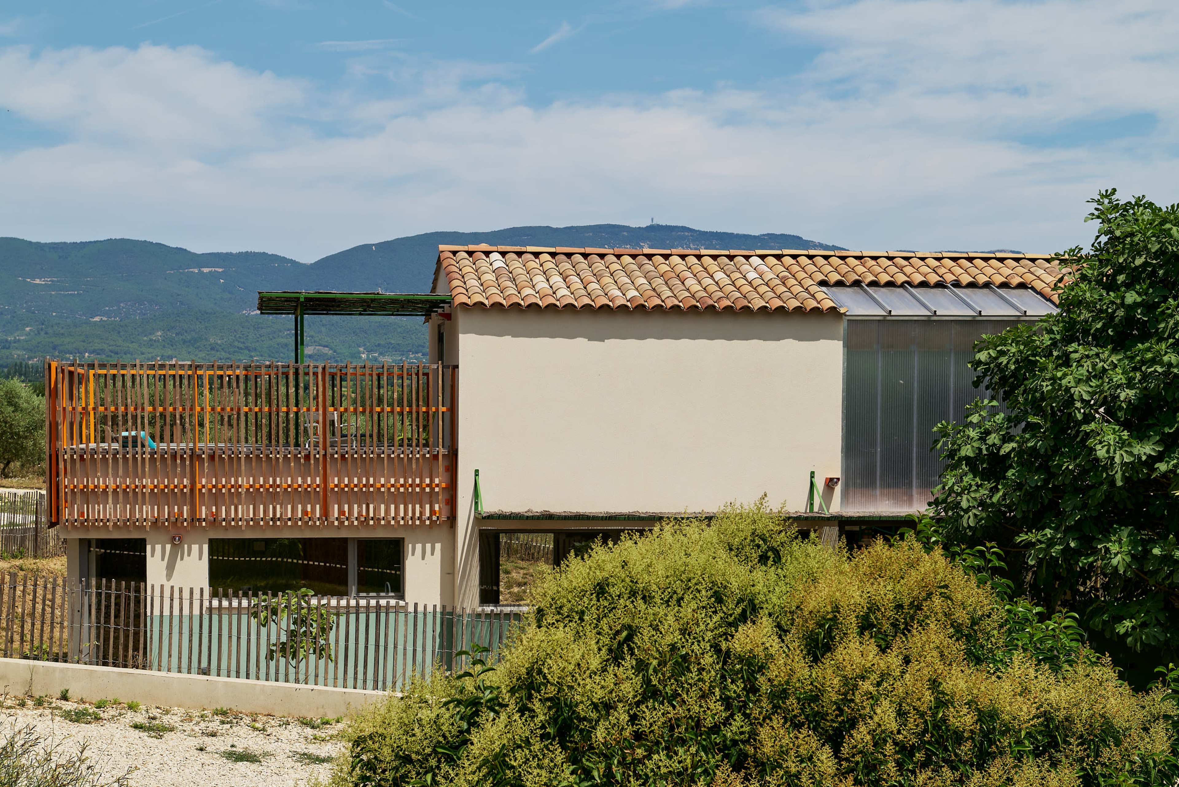A modern two-story house with a tiled roof features a wooden balcony and large glass windows, surrounded by greenery and mountain views.