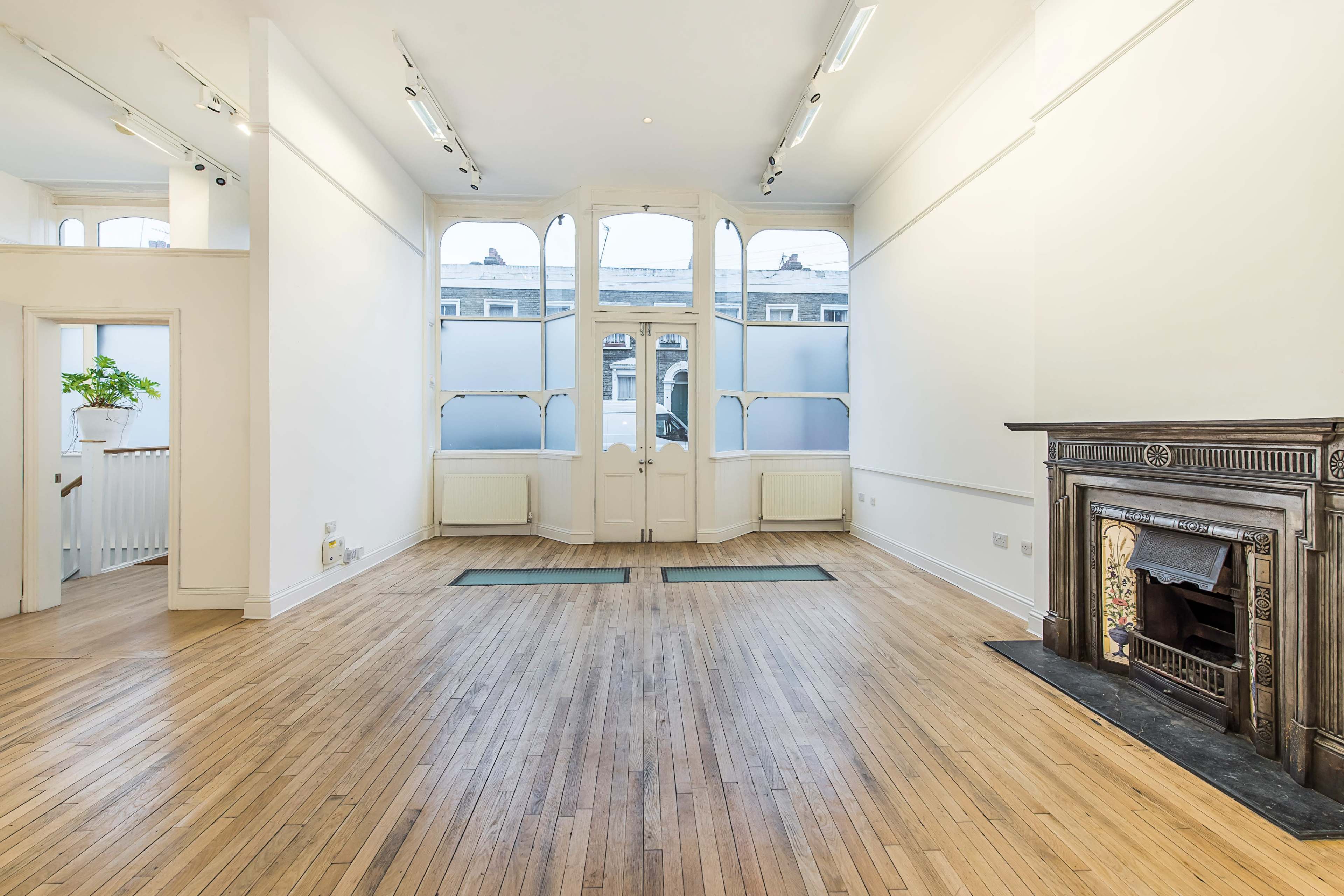 A spacious, empty room with wooden flooring, large windows, and an ornate fireplace against one wall.