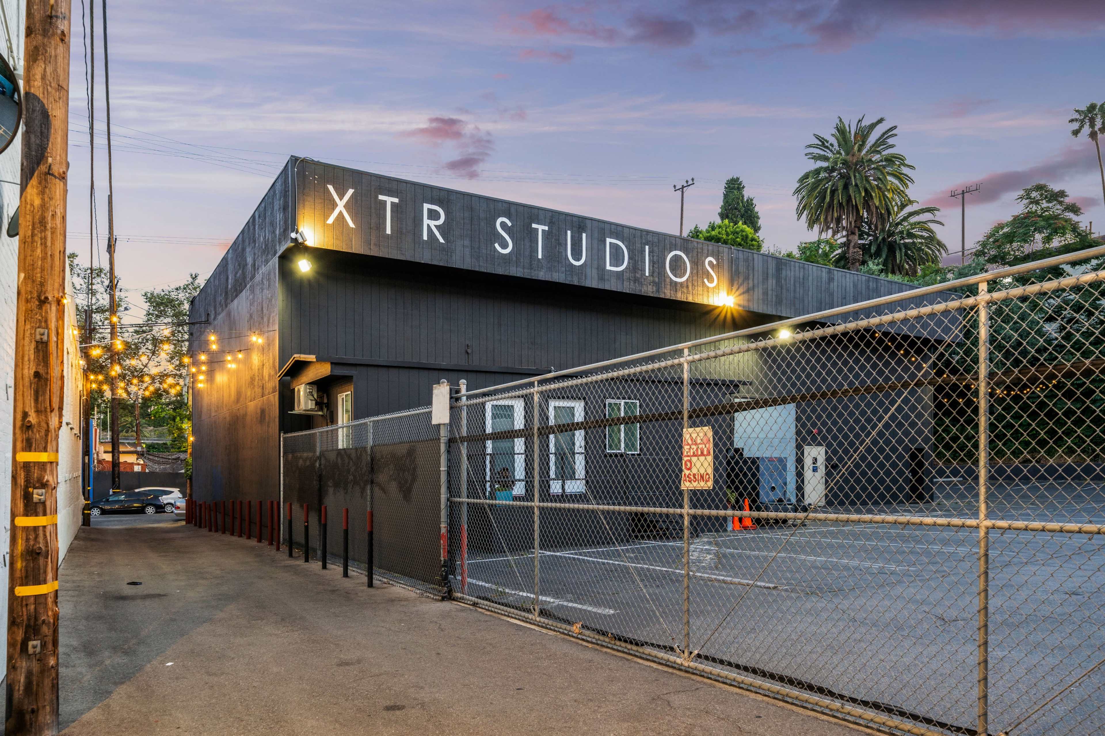 A contemporary building labeled "XTR STUDIOS" surrounded by a chain-link fence, with palm trees and a sunset sky in the background.