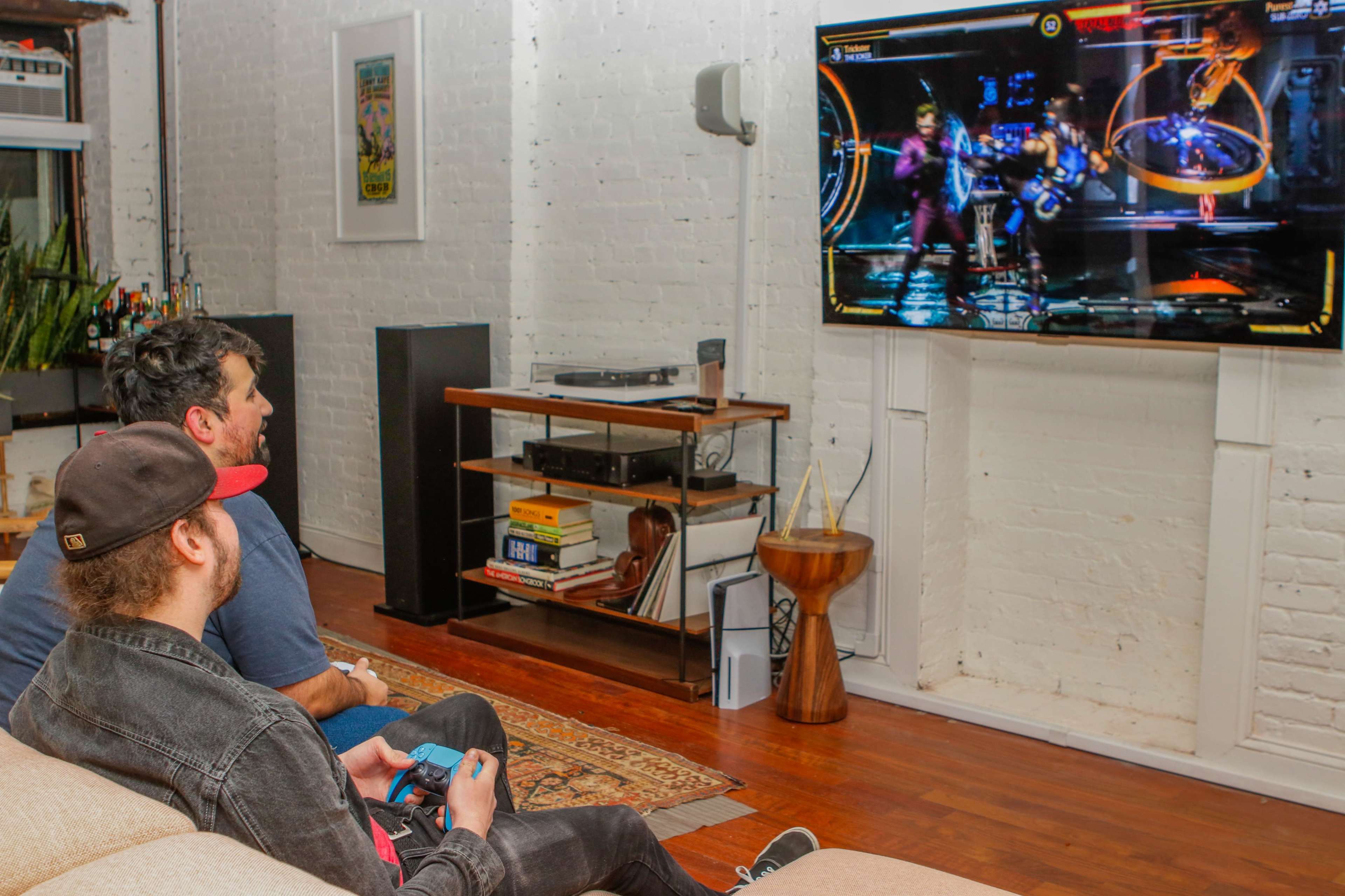 Two men sit on a couch in a living room, playing a video game on a wall-mounted television.