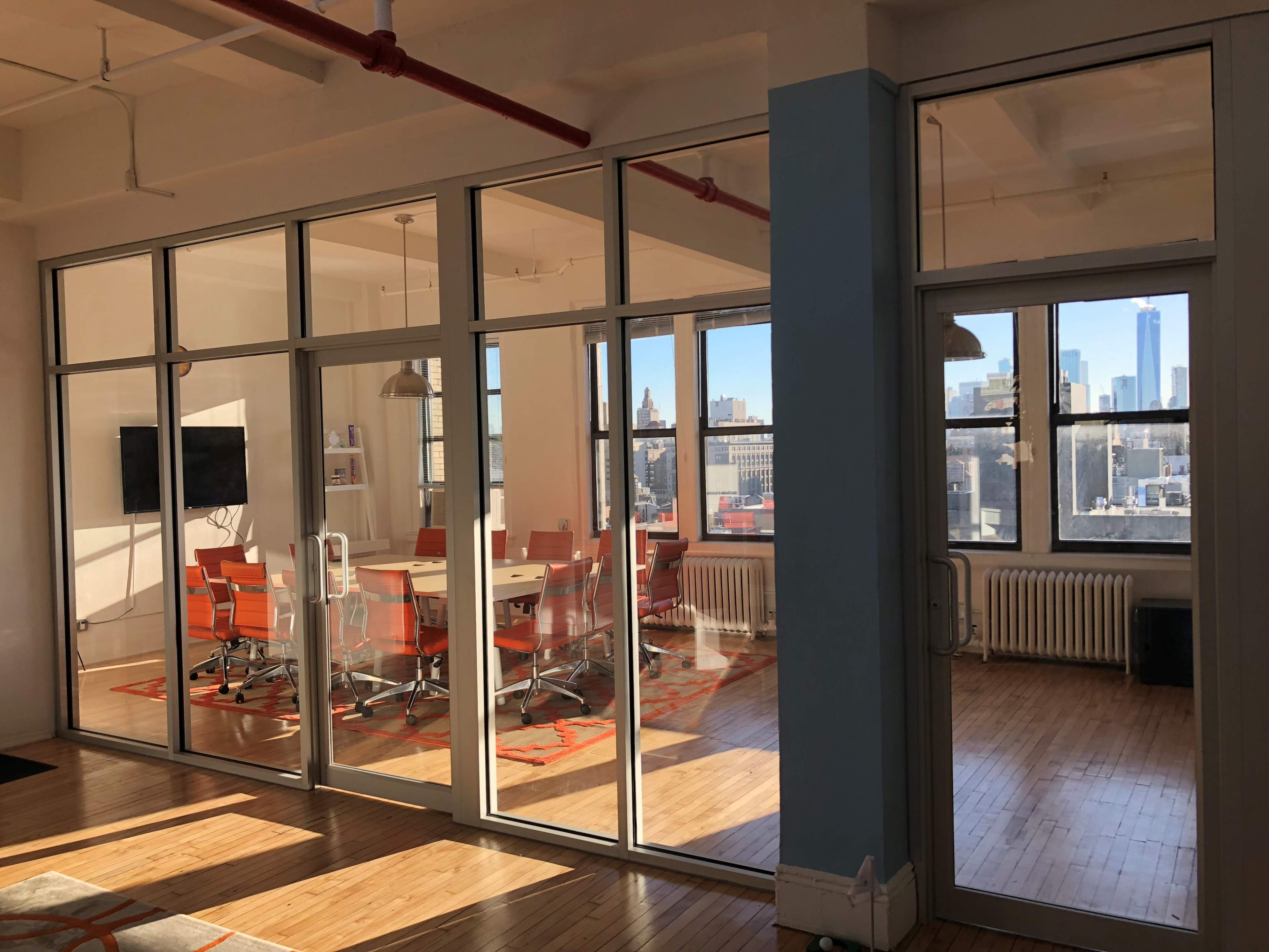 A modern conference room with glass walls, featuring a central table surrounded by orange chairs, with city skyline views through large windows.