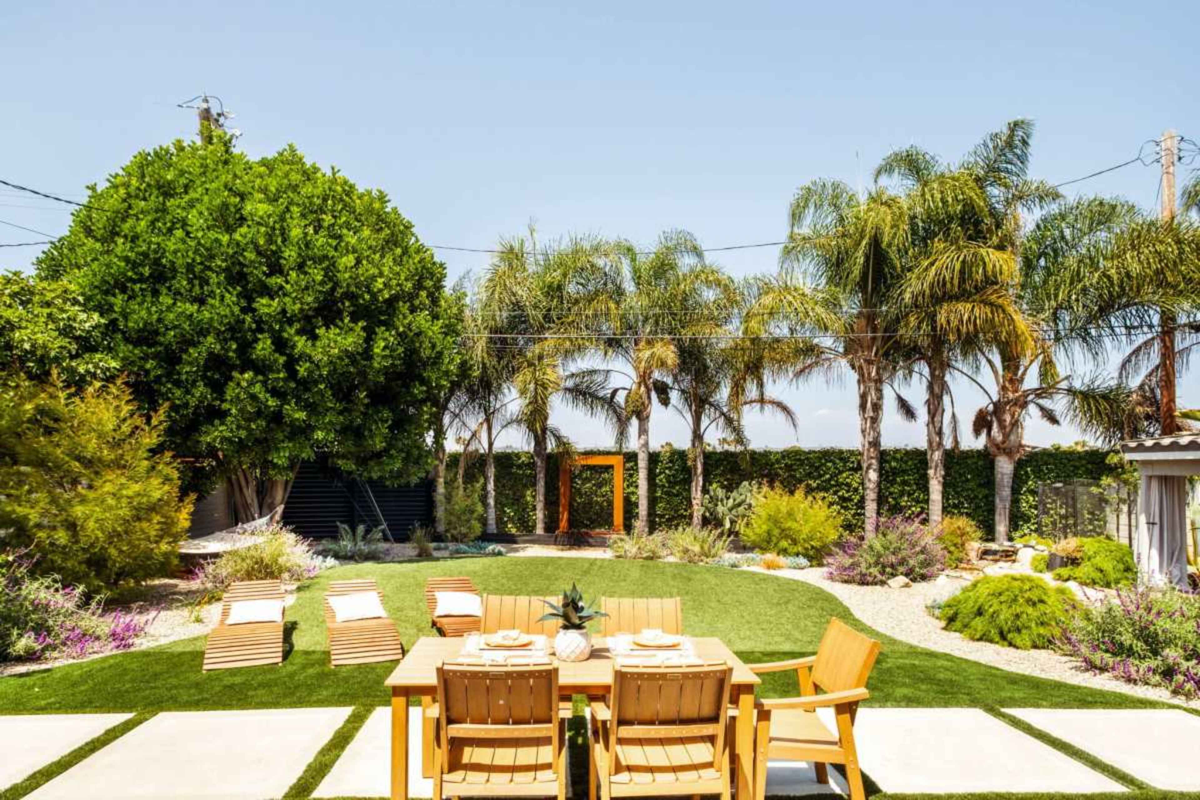 The image shows a backyard with a wooden dining table set for a meal, surrounded by palm trees and landscaped with gravel and various plants.