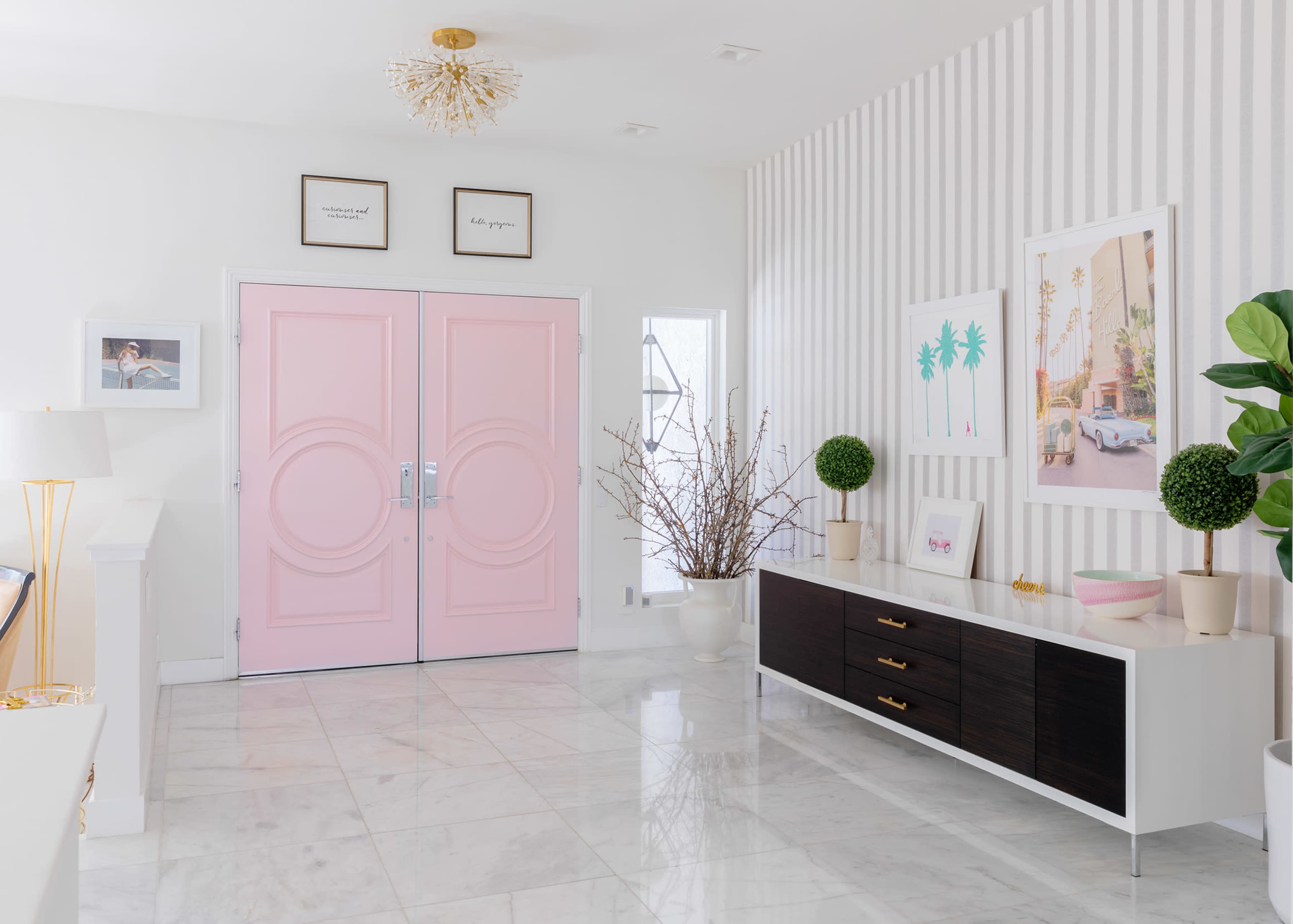 A bright entryway featuring pink double doors, marble flooring, and a modern decor with framed art and decorative plants.