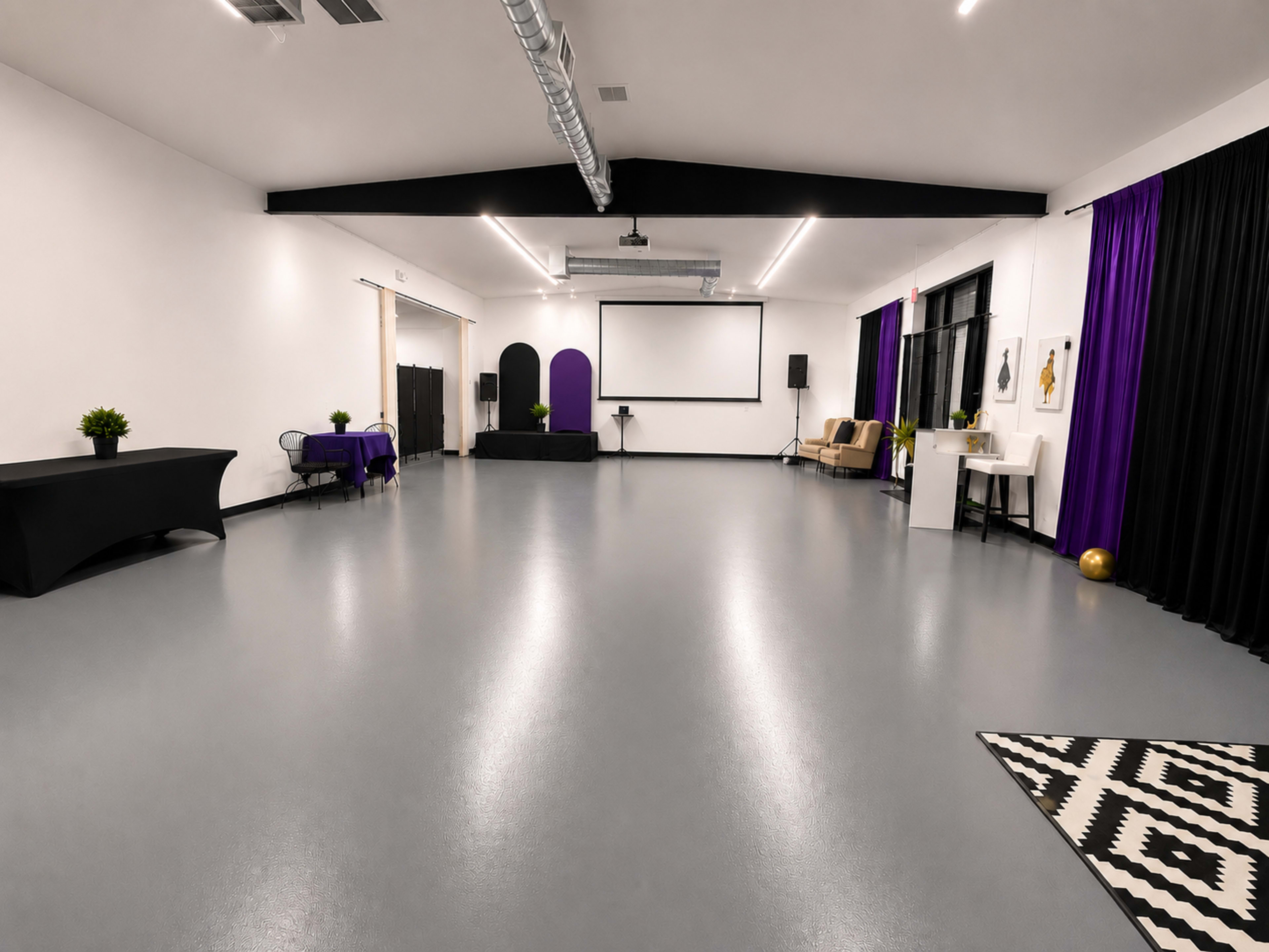 The image shows a spacious, well-lit interior room with a polished gray floor, featuring a large blank screen, a black table, and decorative purple accents.