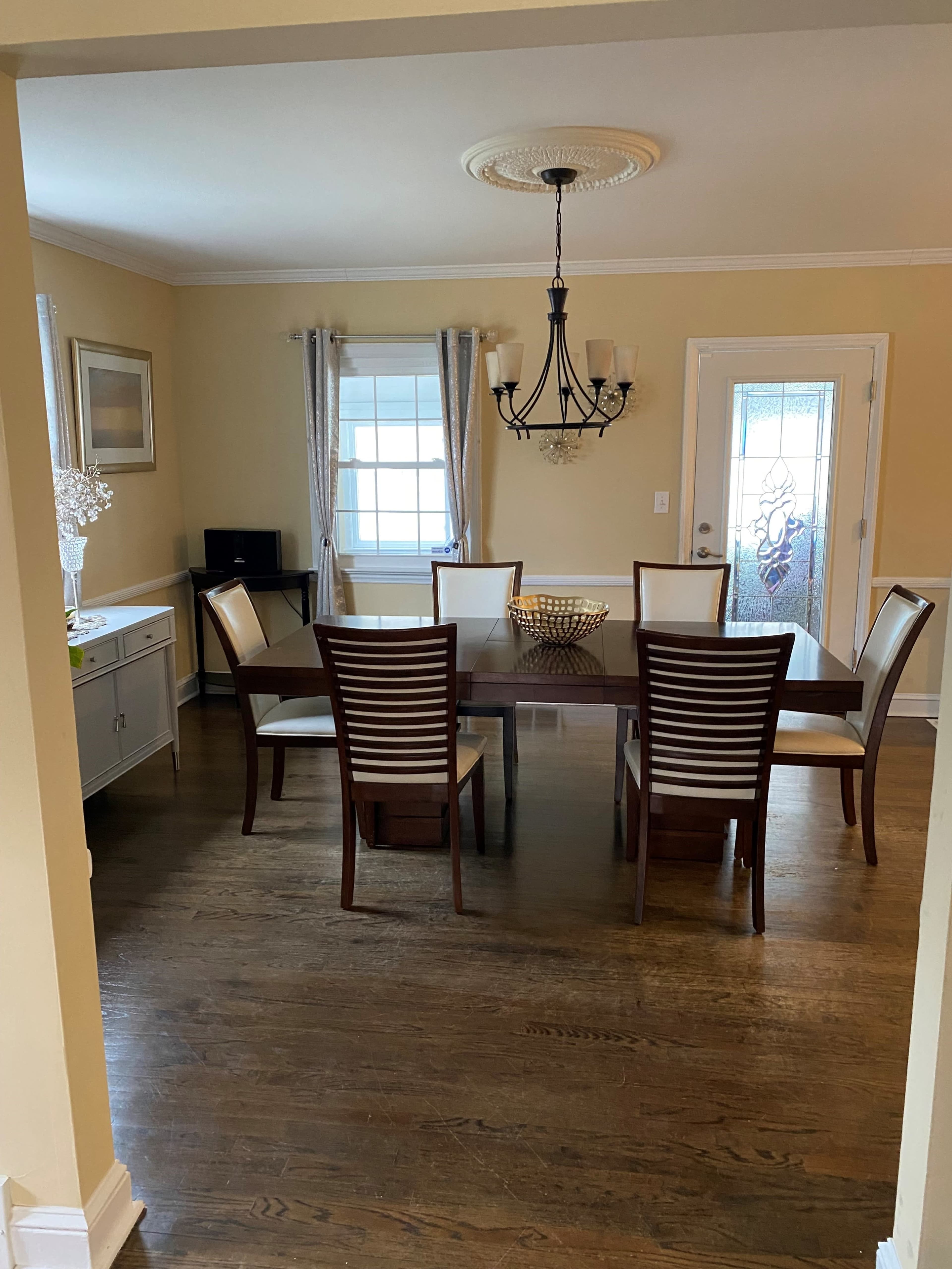 A dining room features a rectangular table surrounded by six wooden chairs, with a chandelier overhead and a sideboard along one wall.