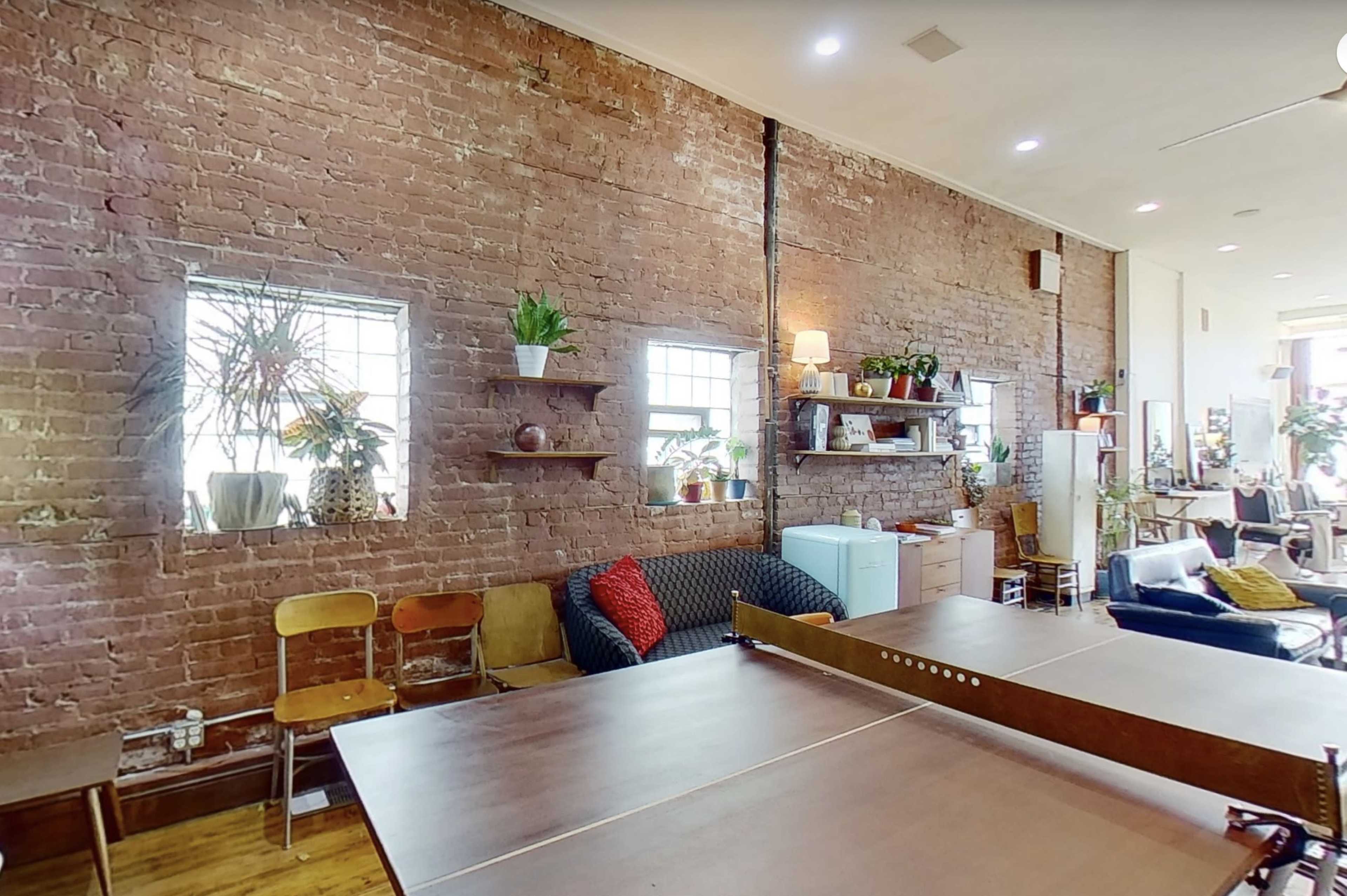 The image shows an interior space with exposed brick walls, two windows, shelves displaying plants, and a table in the foreground.
