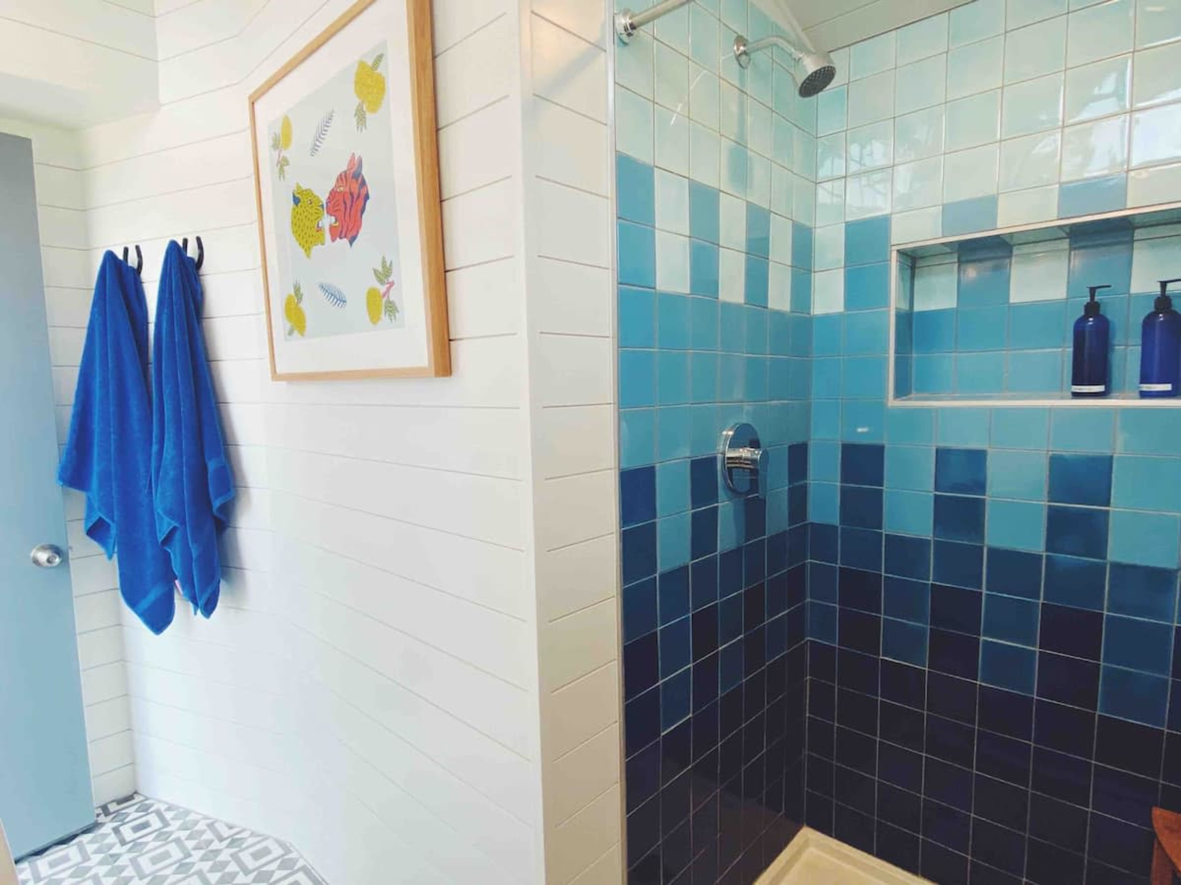 The image shows a modern bathroom with a blue tiled shower area, a framed floral artwork on the wall, and two blue towels hanging on a hook.