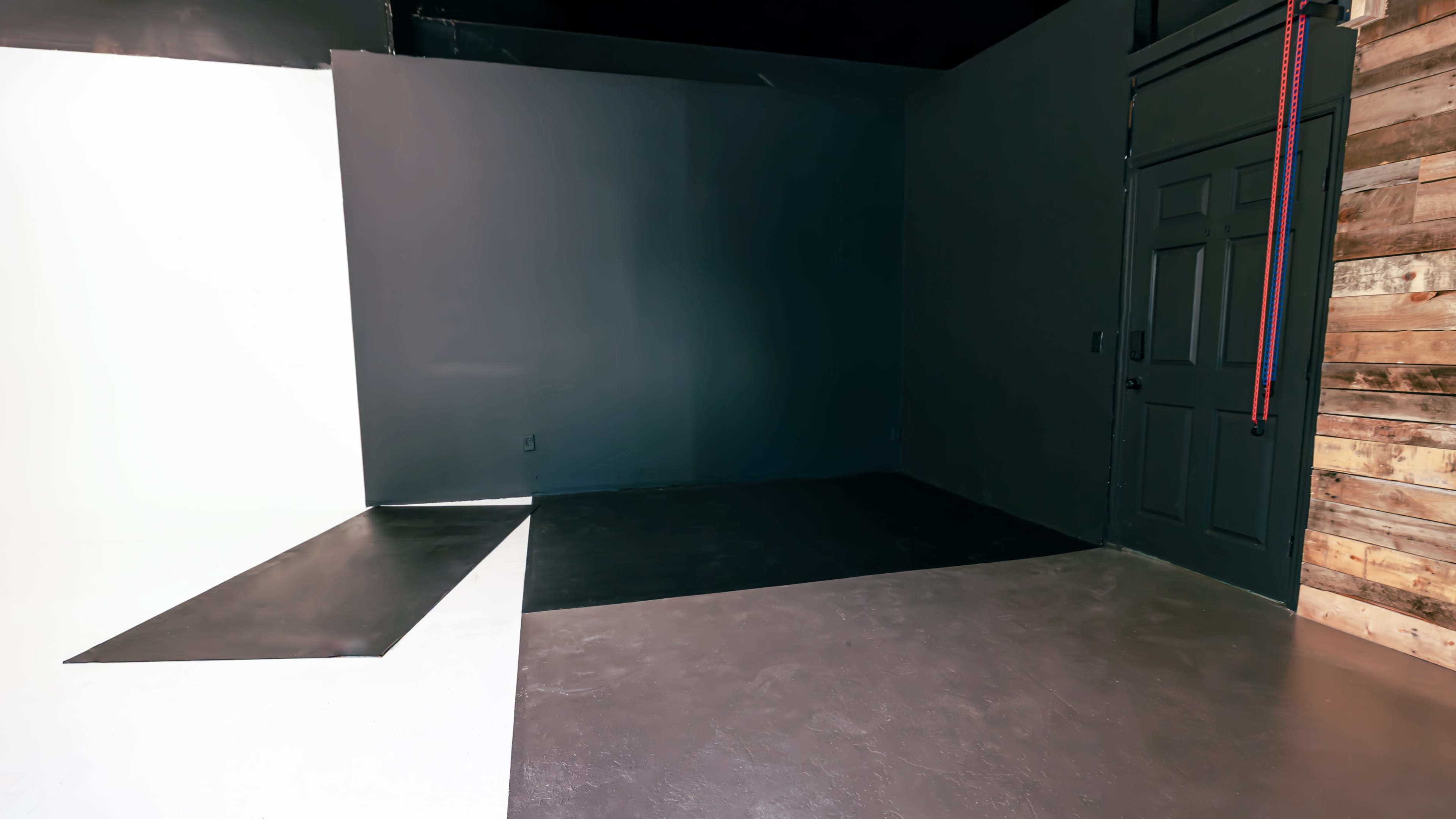 The image shows a sparsely furnished room with gray walls, a black floor, and a white corner contrasting with the darker surfaces.