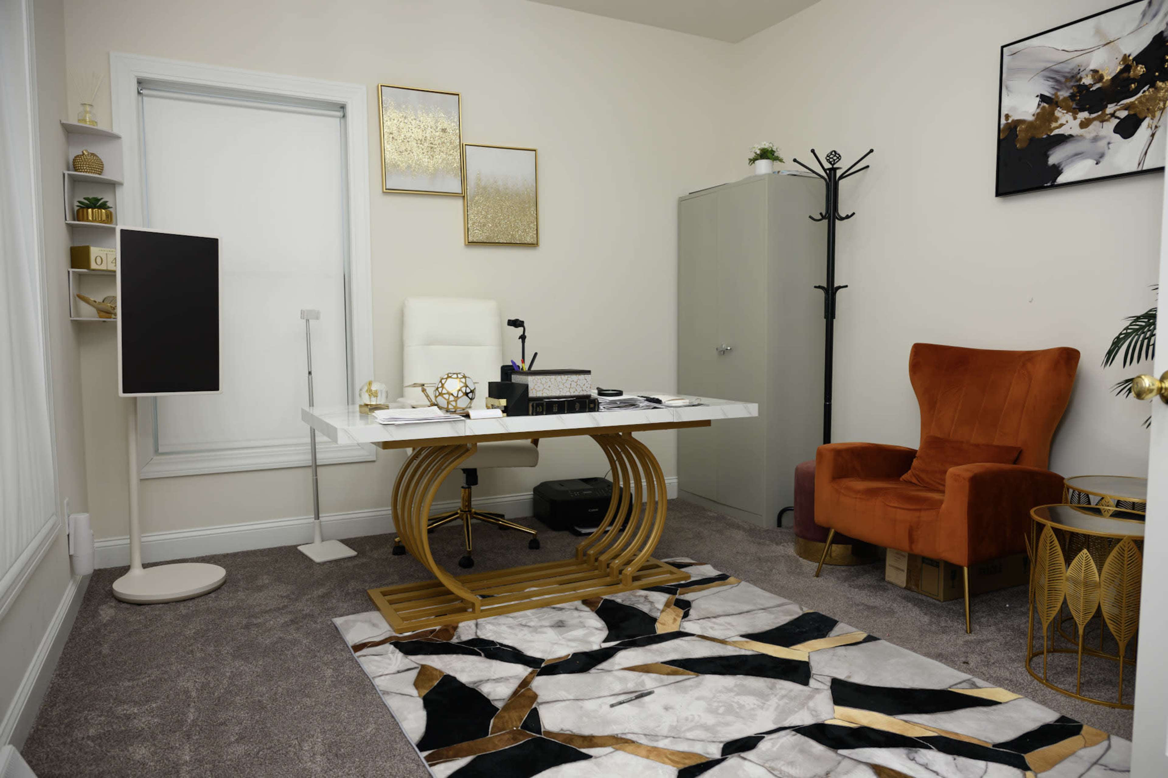 The office features a modern desk with a sleek design, an orange armchair, and decorative wall art, all set against a neutral-colored wall and plush carpet.