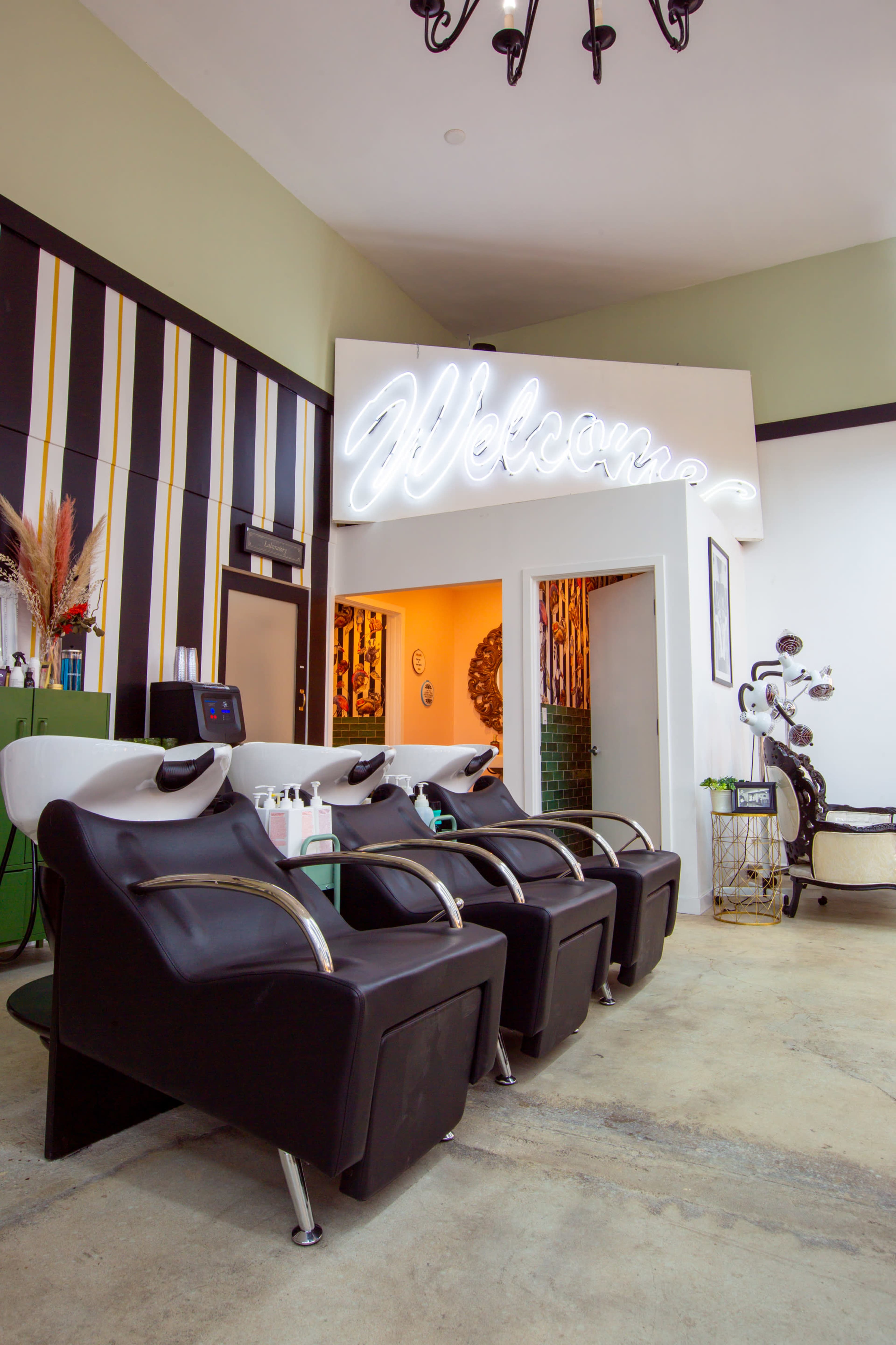 A modern hair salon interior with three black shampoo chairs arranged in front of a illuminated "Welcome" sign.