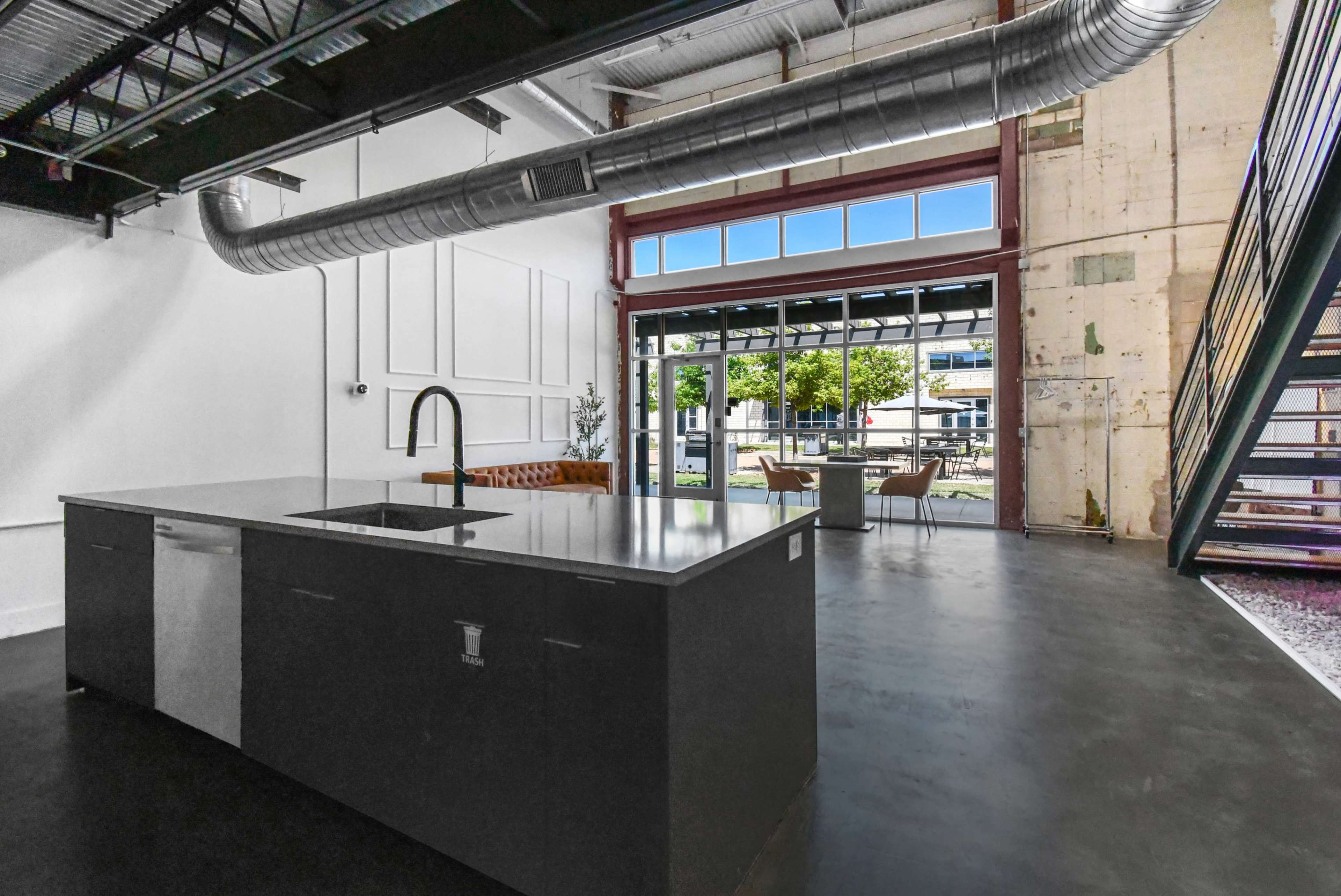 The image shows a modern kitchen with a central island, large windows allowing natural light, and a view of an outdoor seating area.