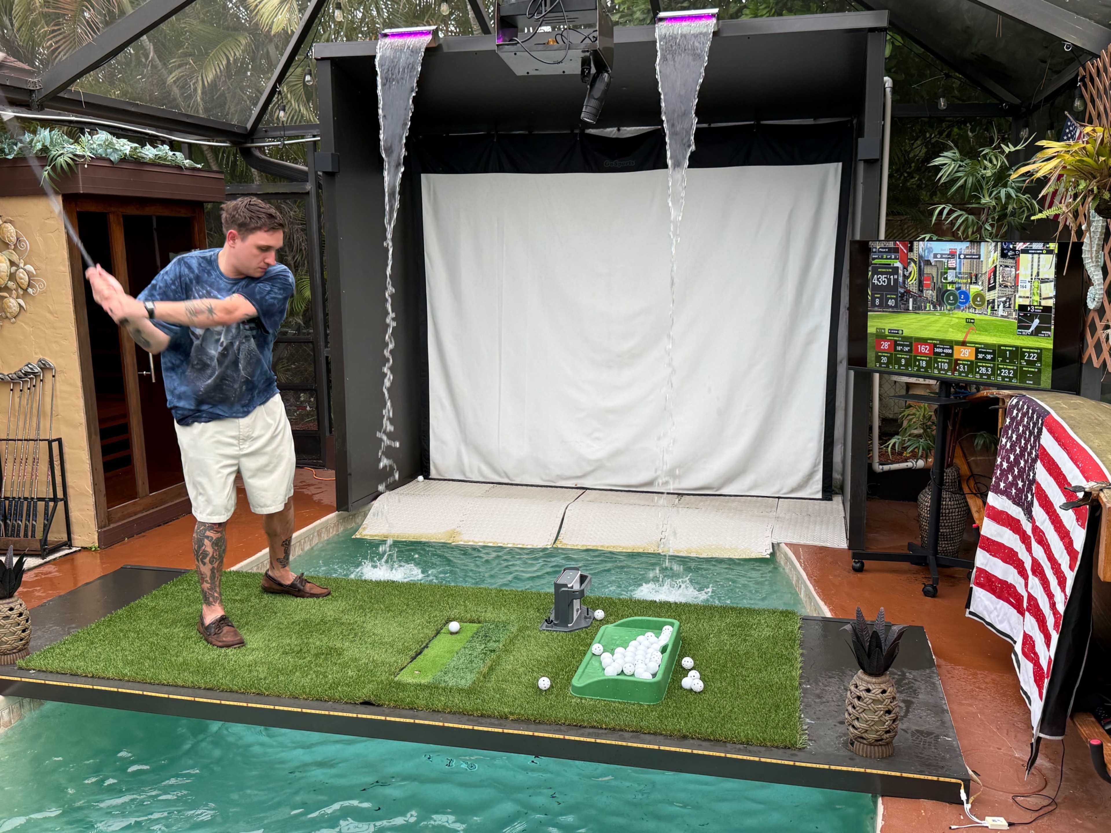 A man is swinging a golf club in an indoor simulated driving range equipped with a screen and a water feature, while a basket of golf balls is on a green surface nearby.