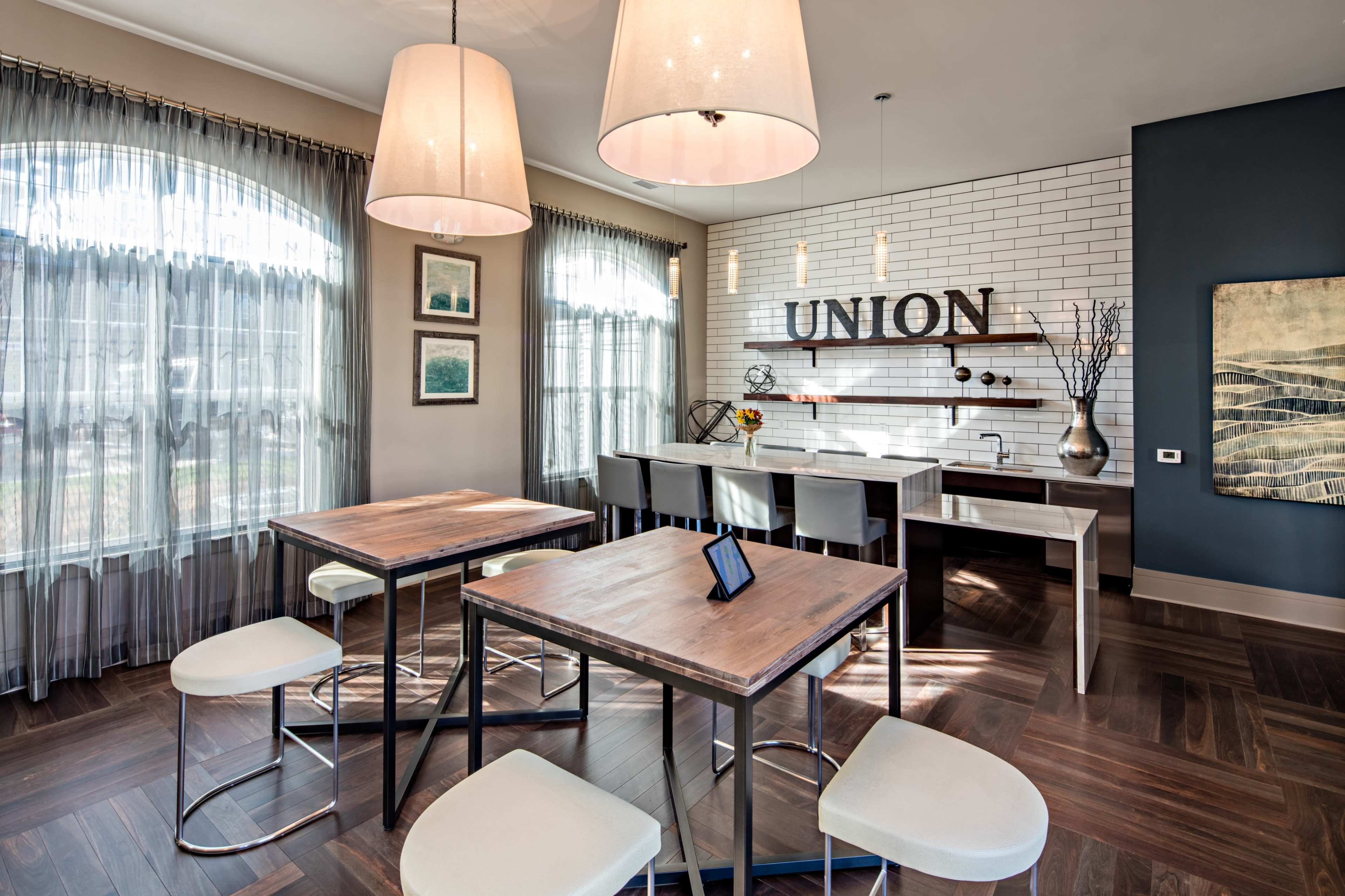 A modern dining area with two square tables, white bar stools, large pendant lights, and a wall featuring the word "UNION" in bold letters.