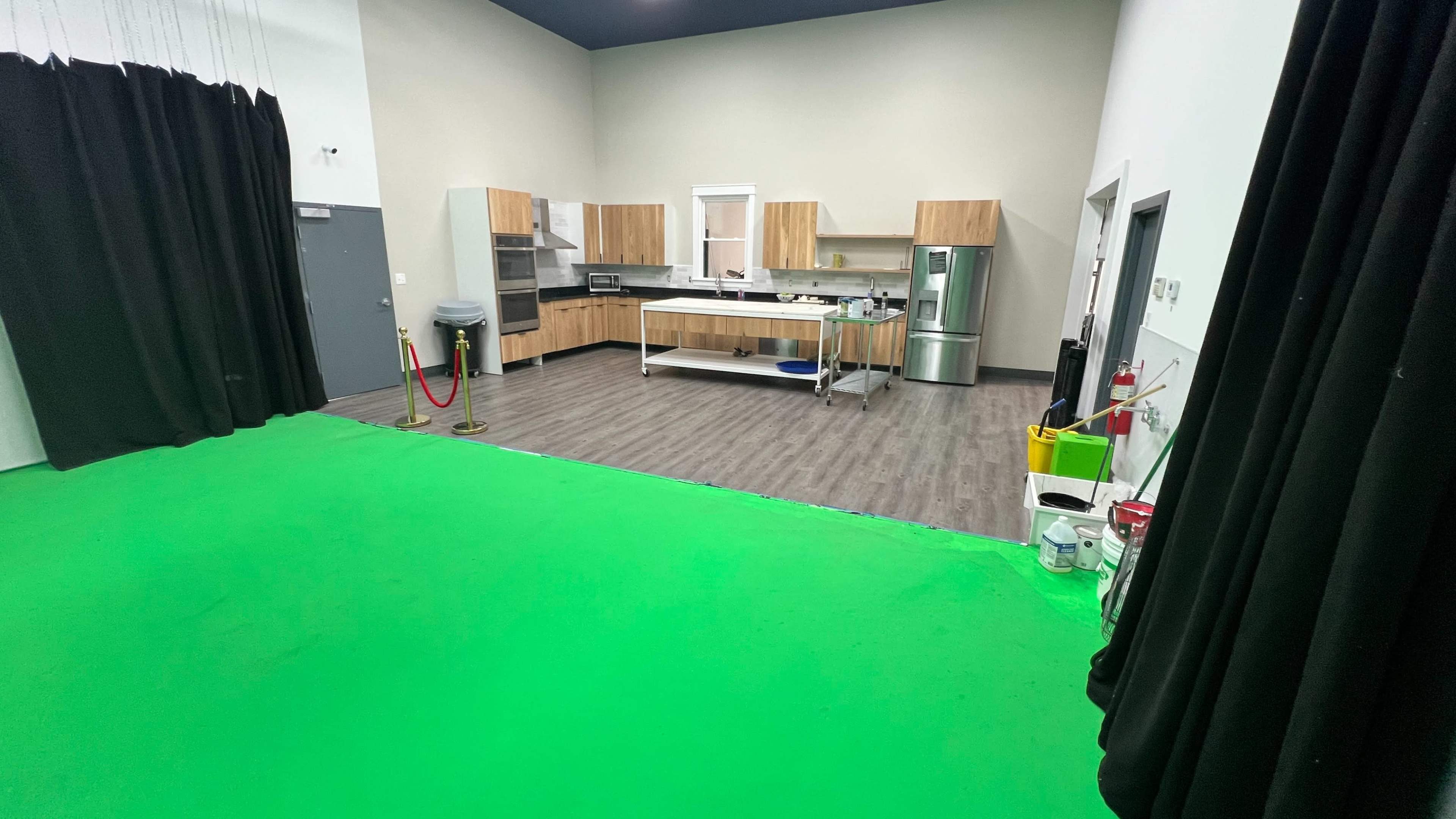 The image shows a room with a green floor, a modern kitchen setup on one side, and black curtains lining the walls.