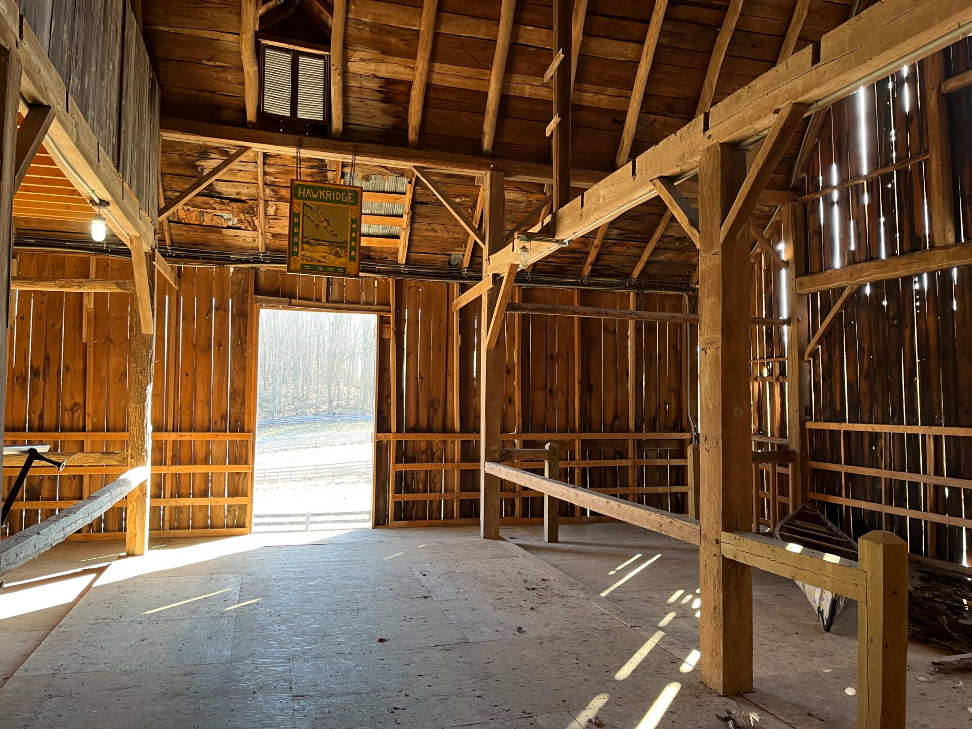 The interior of a wooden barn features exposed beams, a light source from the open door, and wooden railings along the perimeter.