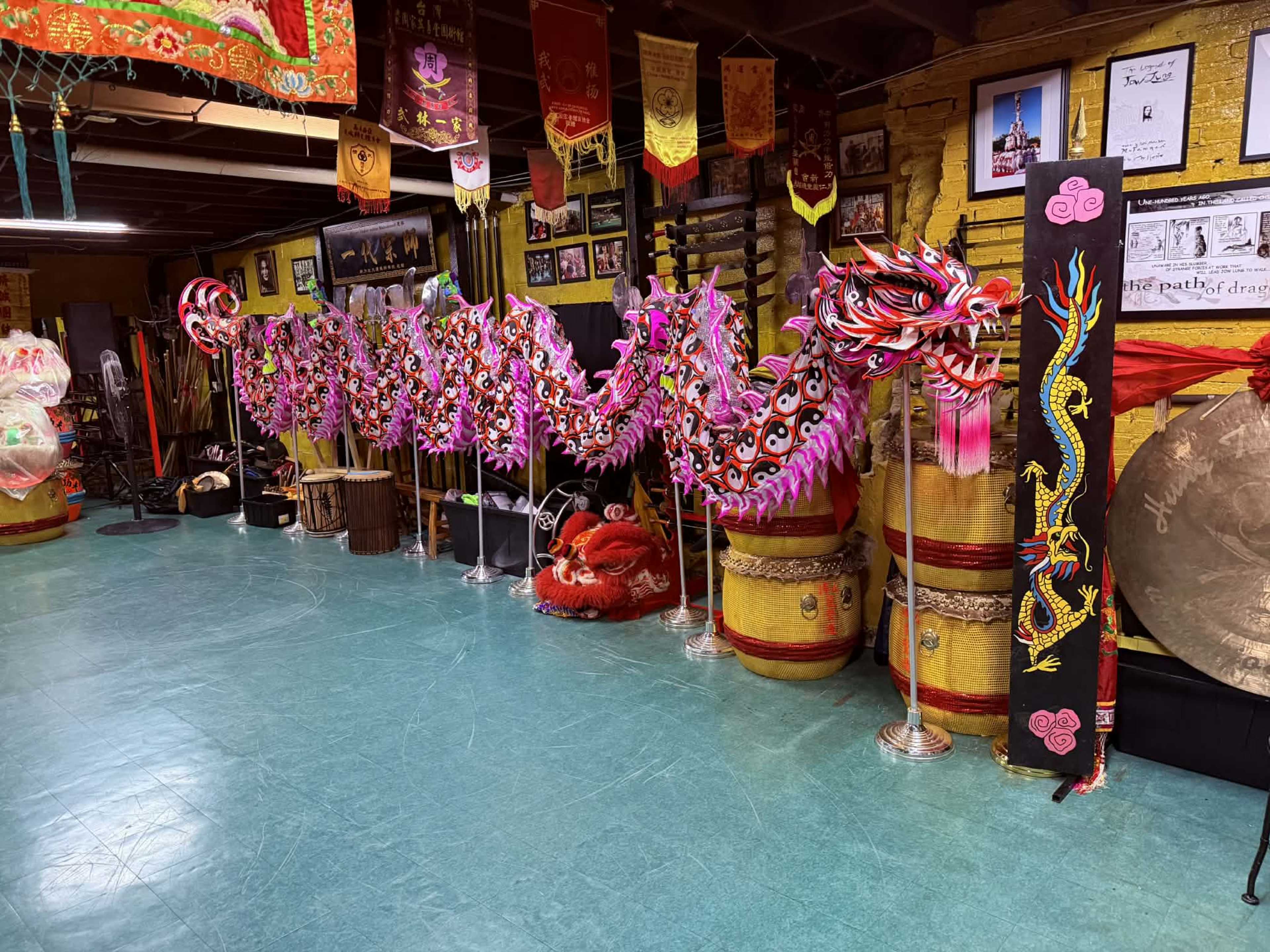 The image shows a decorative dragon dance display, featuring colorful dragon costumes and traditional instruments, arranged in a dimly lit room adorned with banners and photographs.