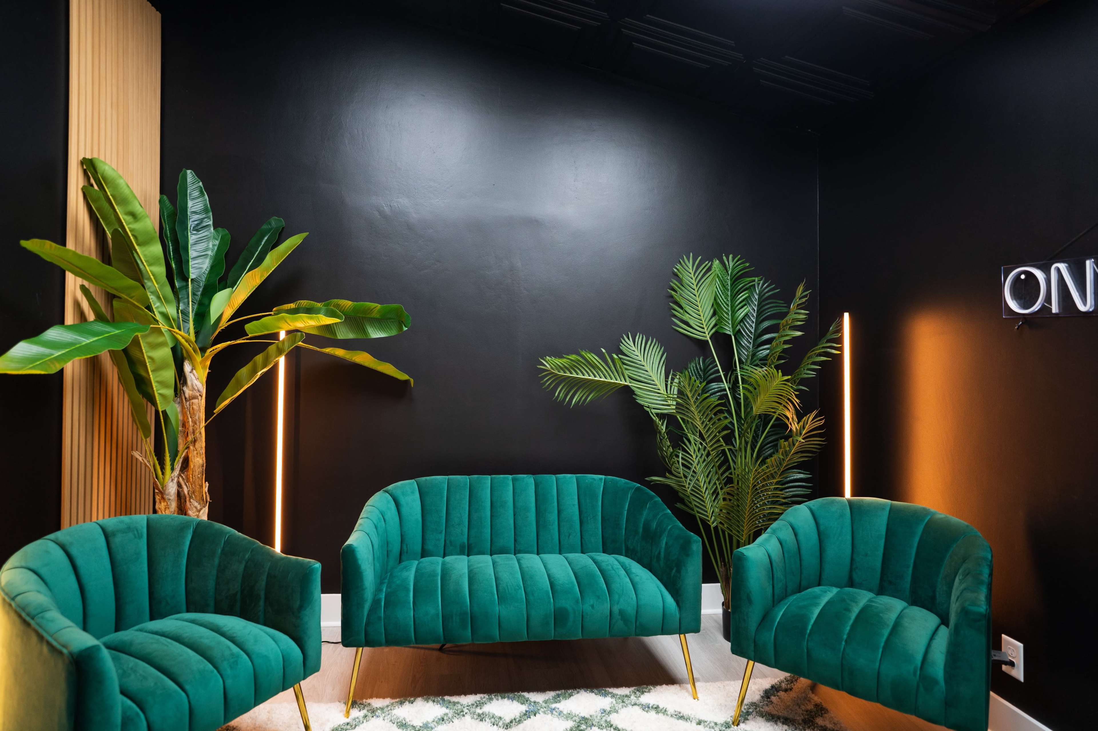 The image features a modern seating area with three green upholstered sofas arranged around a decorative rug, complemented by indoor plants against a black wall.
