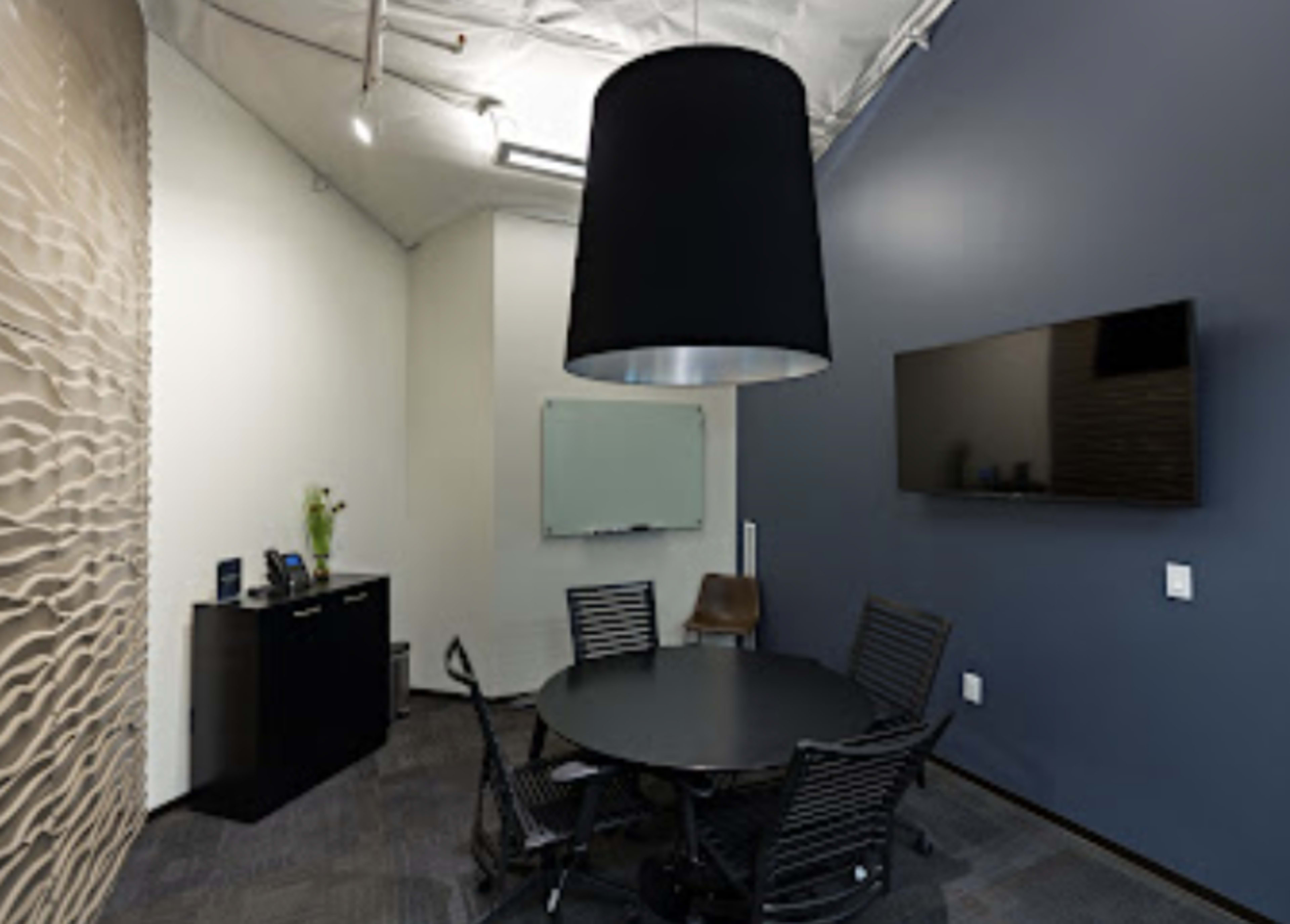The image shows a modern conference room featuring a round black table surrounded by six chairs, a large pendant light, and a wall-mounted television.