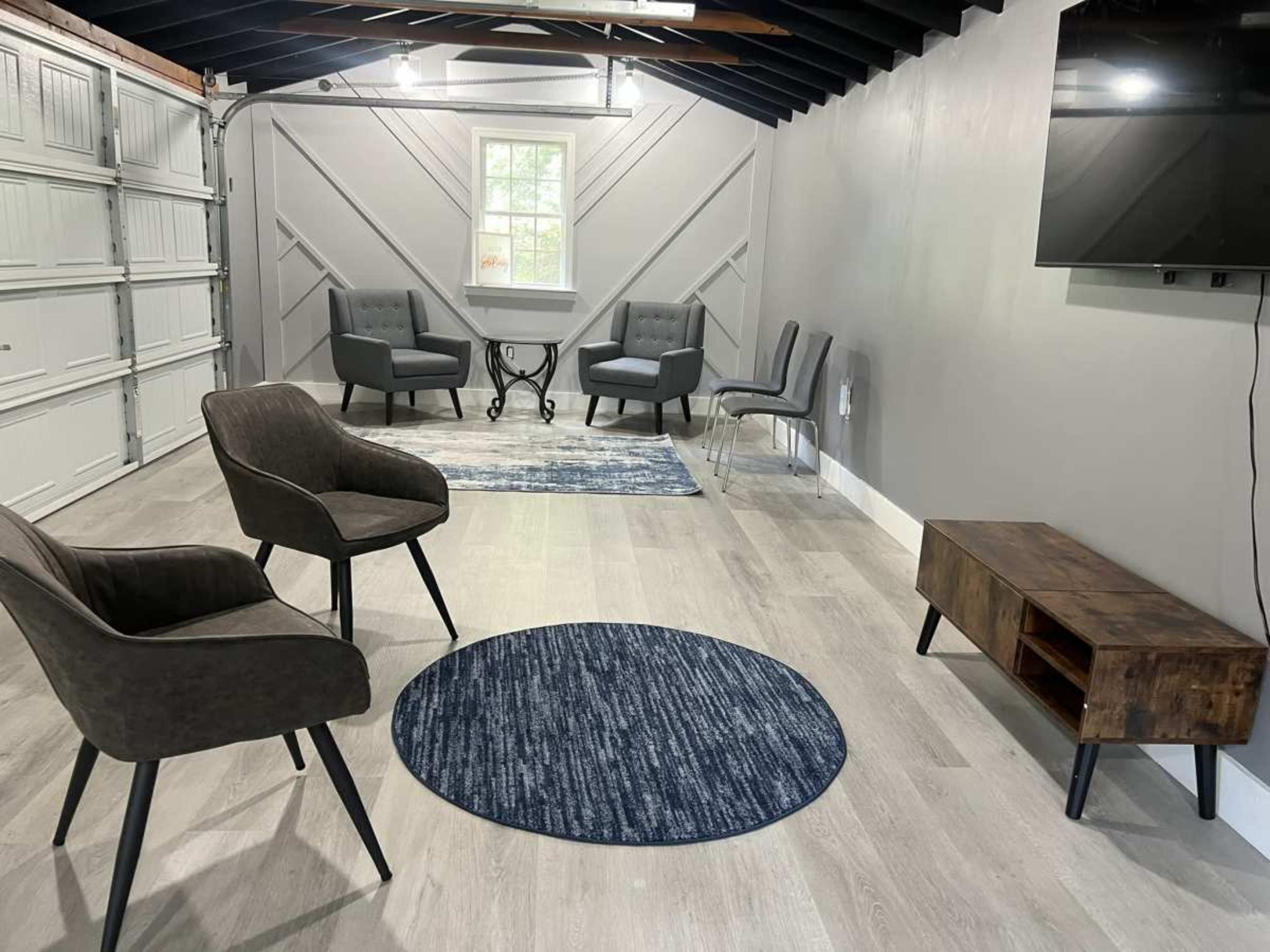 The image shows a sparsely furnished interior space with gray walls, a wooden ceiling, and several seating arrangements, including two dark chairs and a wooden media console.