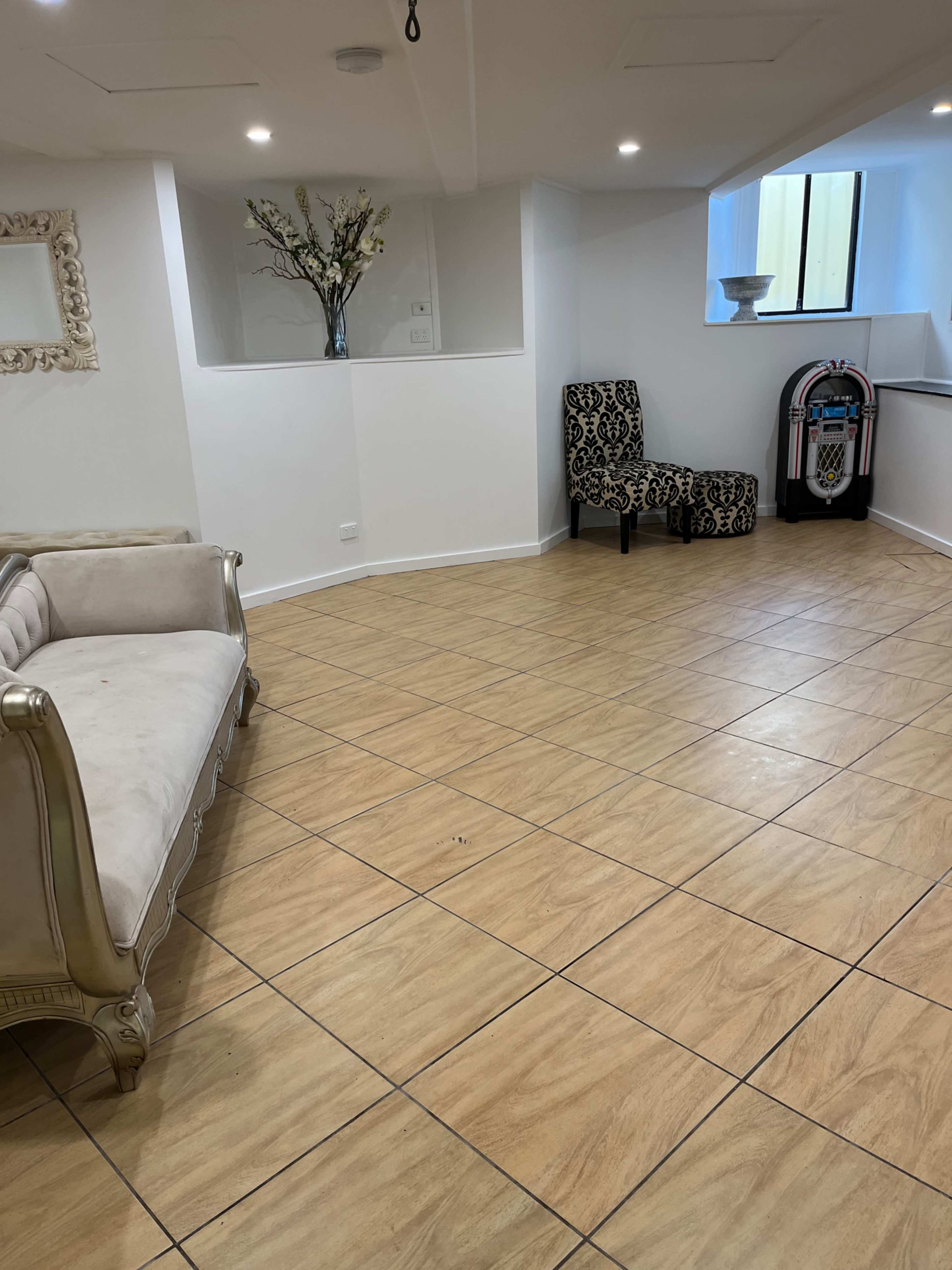 The image shows a spacious, well-lit room with tan tiled flooring, a decorative floral arrangement, a ornate white-framed mirror, and two patterned chairs beside a jukebox.