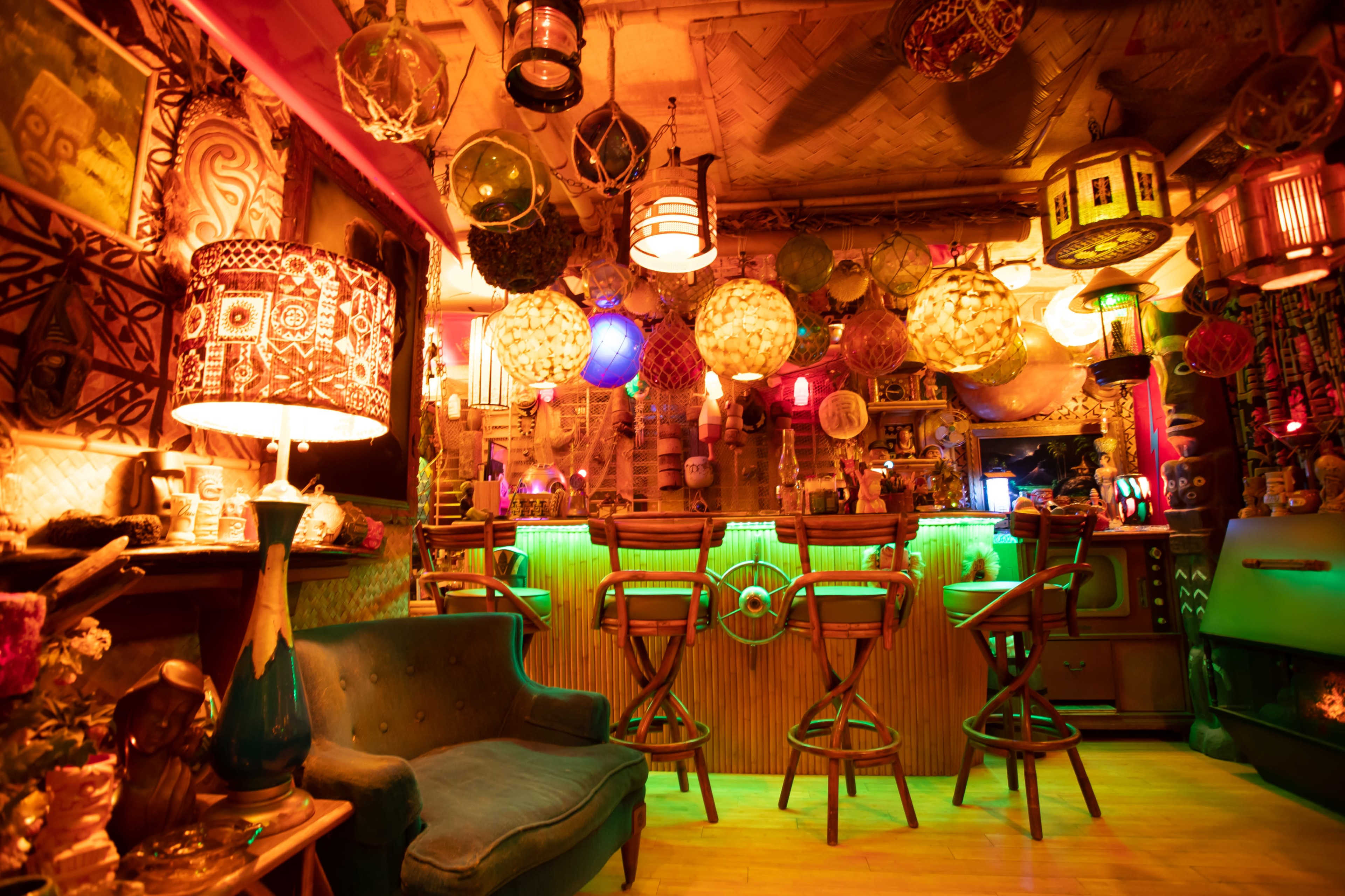 A cozy bar interior features a variety of decorative lanterns and a green-lit counter, with wooden bar stools and a vintage couch.