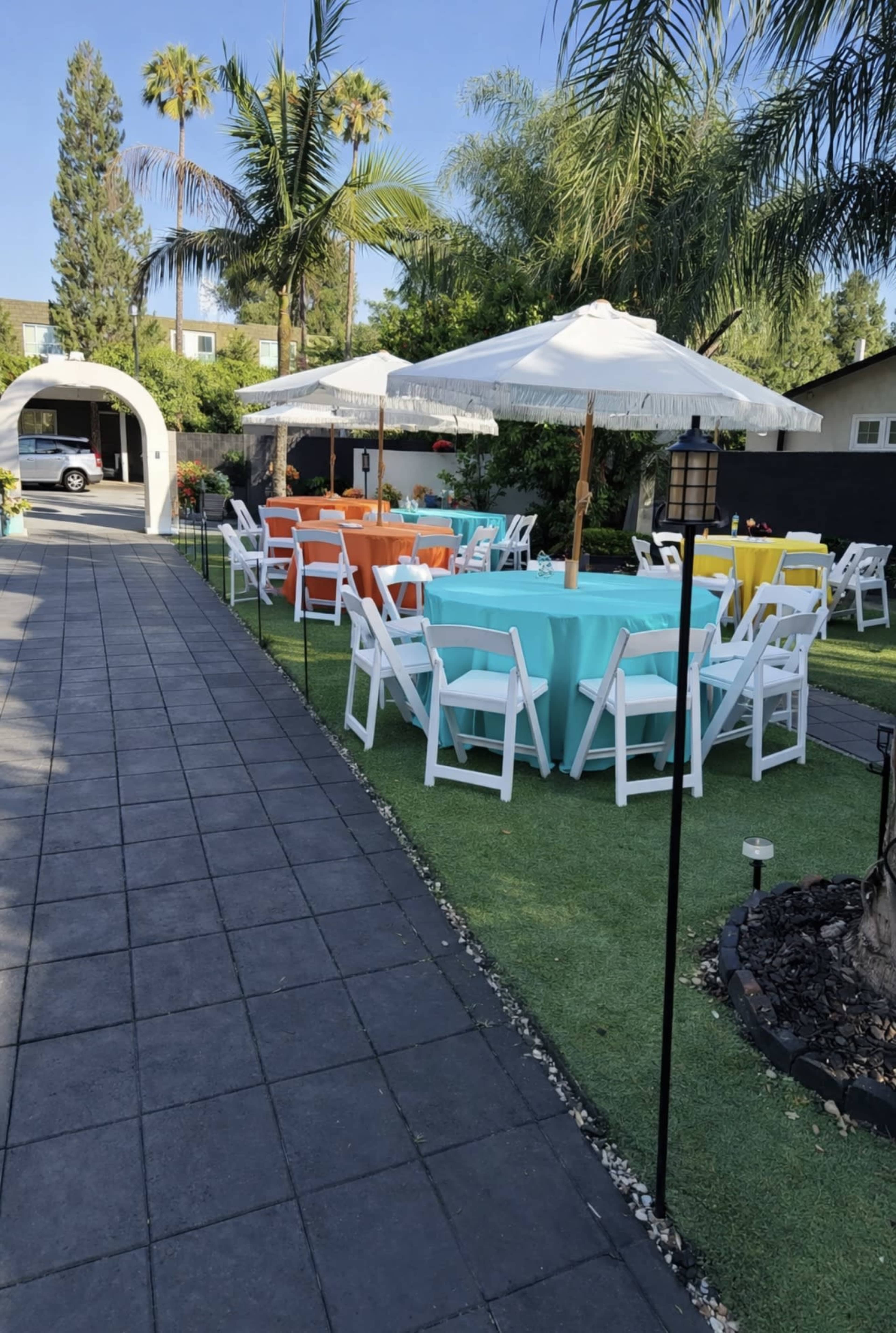 The scene features brightly colored tables with white chairs arranged on a lawn under umbrellas, surrounded by palm trees and a pathway leading to a nearby structure.