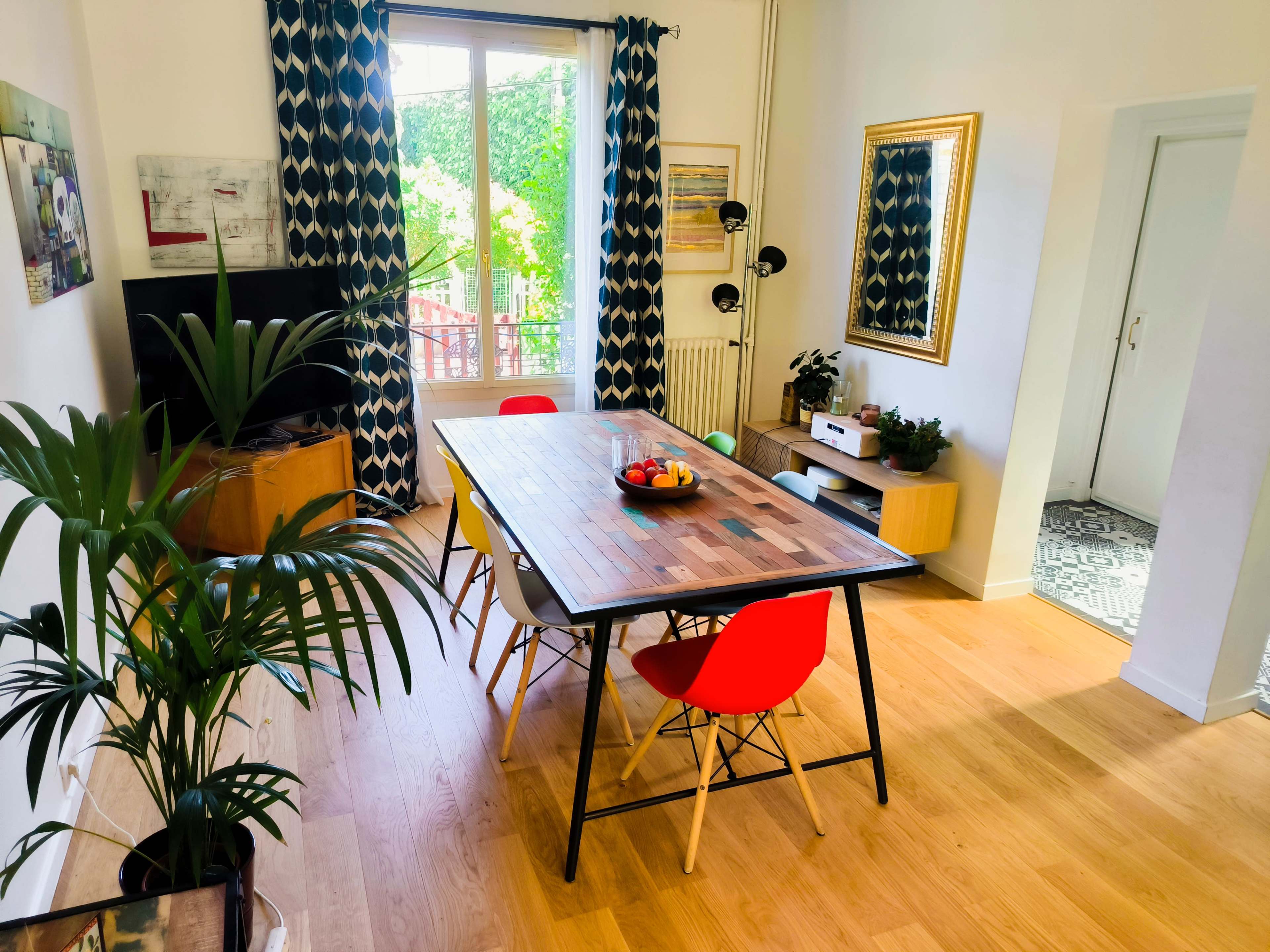 A dining area features a wooden table surrounded by colorful chairs, with large windows allowing natural light to fill the space.
