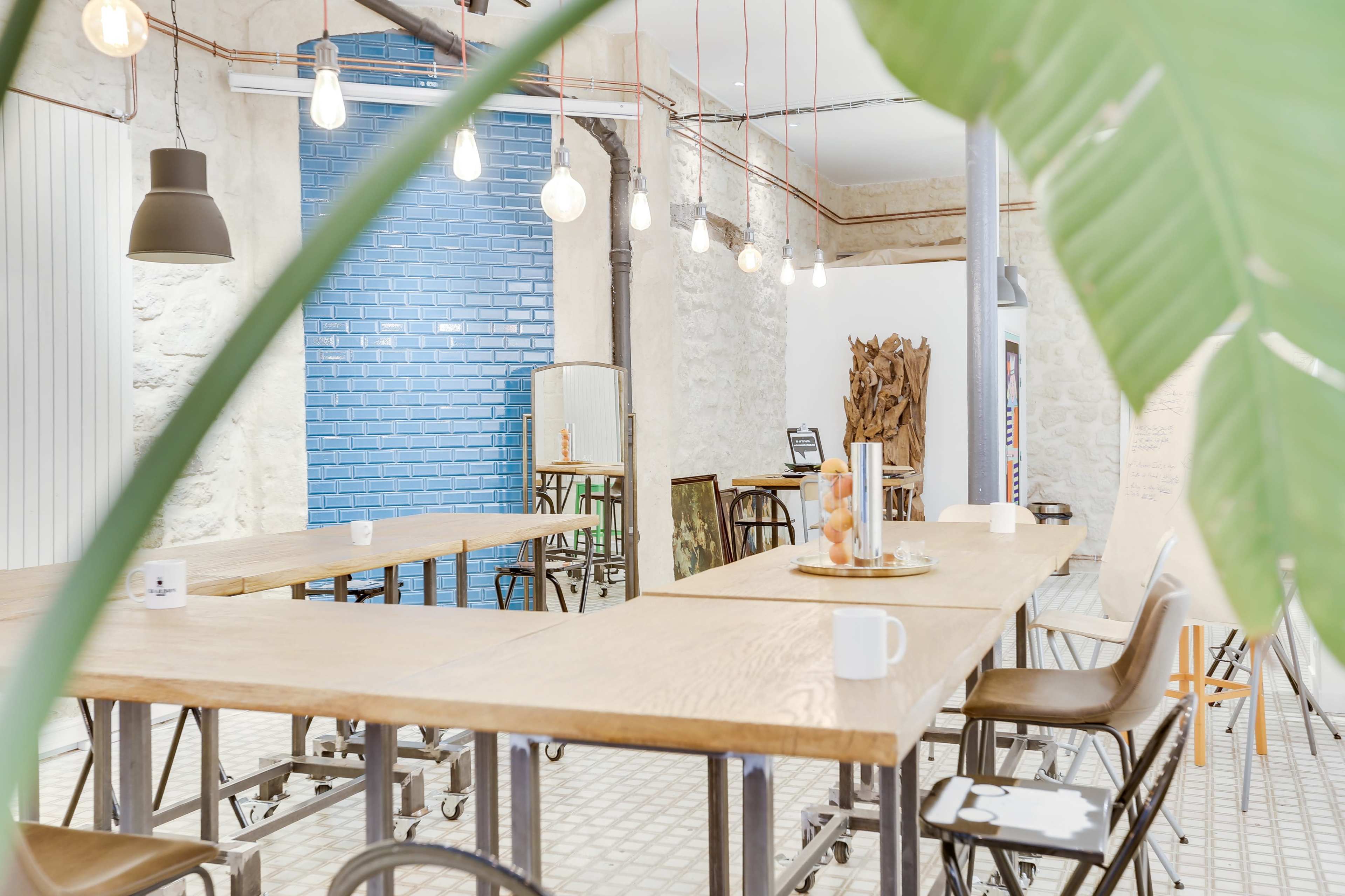 A spacious, modern room with wooden tables, chairs, and a blue tiled wall, illuminated by hanging light bulbs.