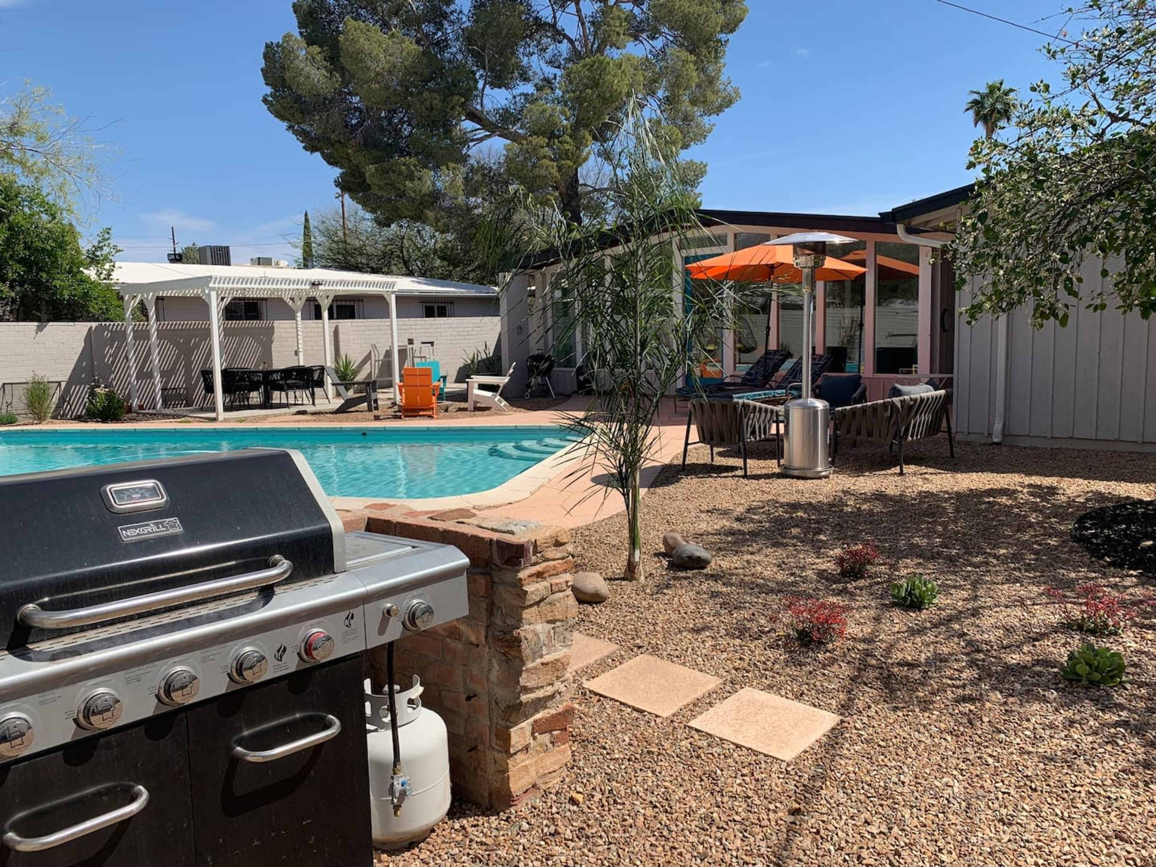 A backyard with a swimming pool, a barbecue grill, lounge chairs, and a covered seating area surrounded by gravel and greenery.