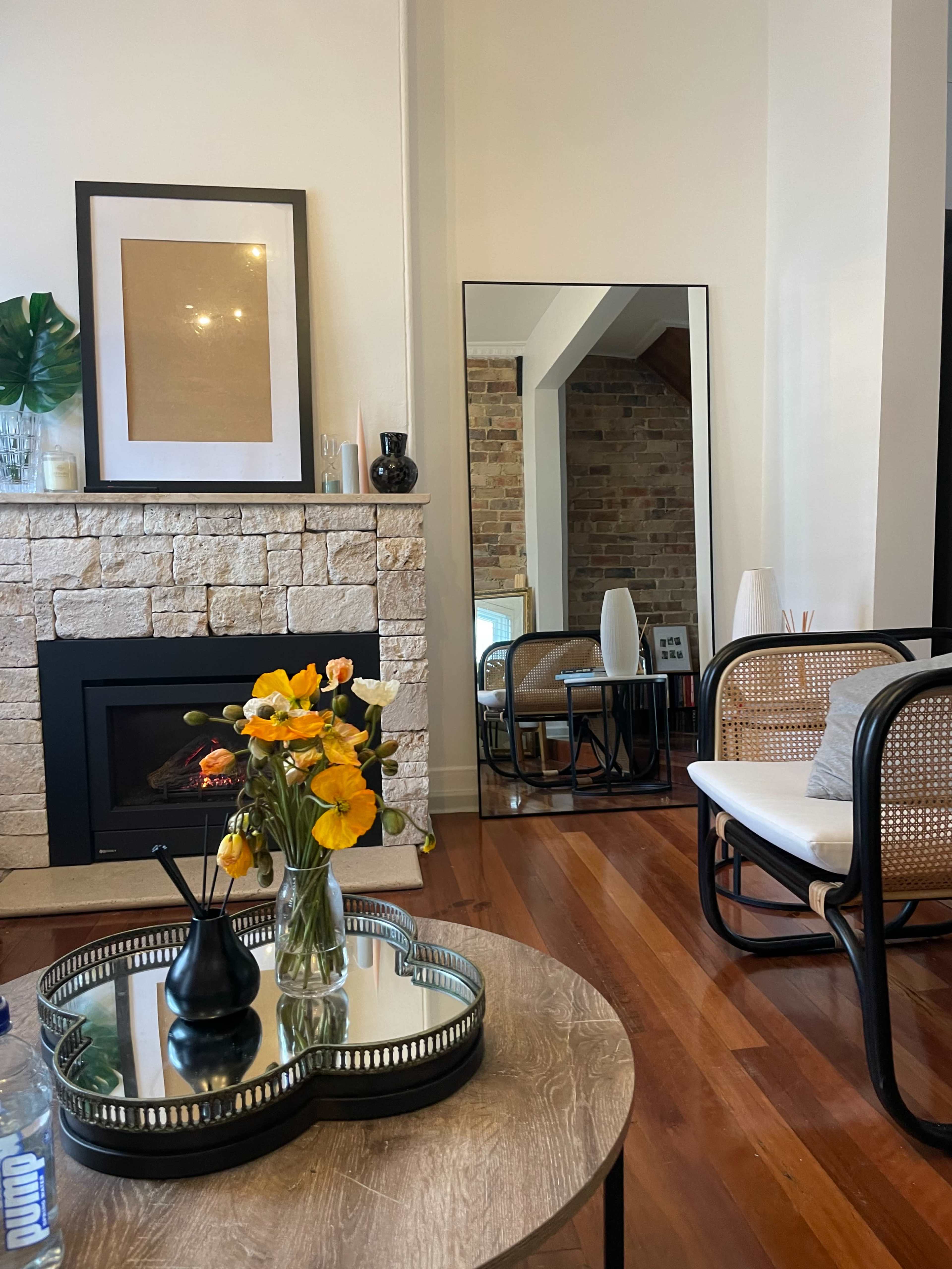 A cozy living room featuring a stone fireplace, a large mirror, a wooden table with a vase of flowers, and modern seating.