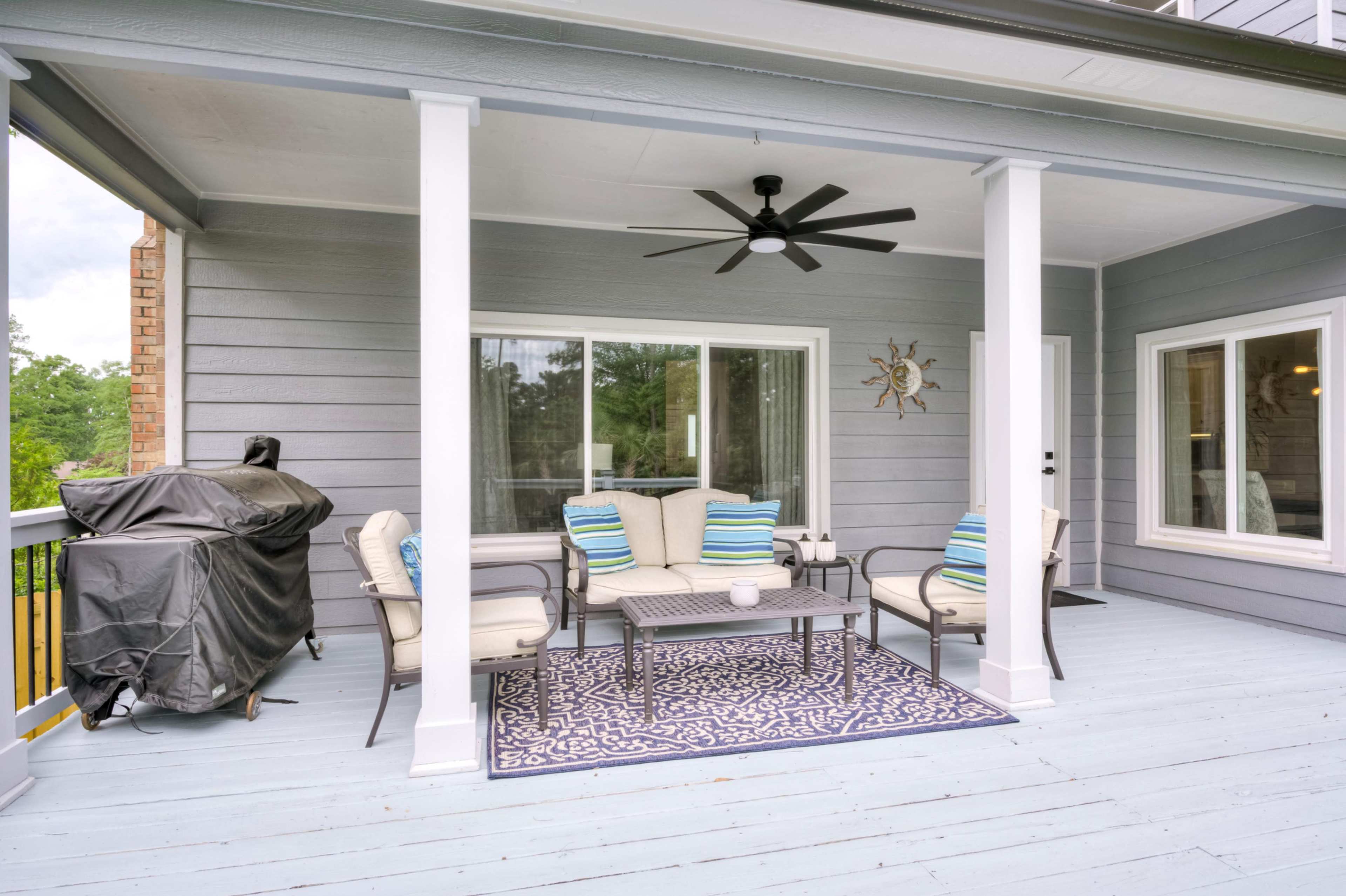 The image shows a covered porch with outdoor furniture, including a seating area with cushions, a coffee table, a fan, and a grill in the corner.