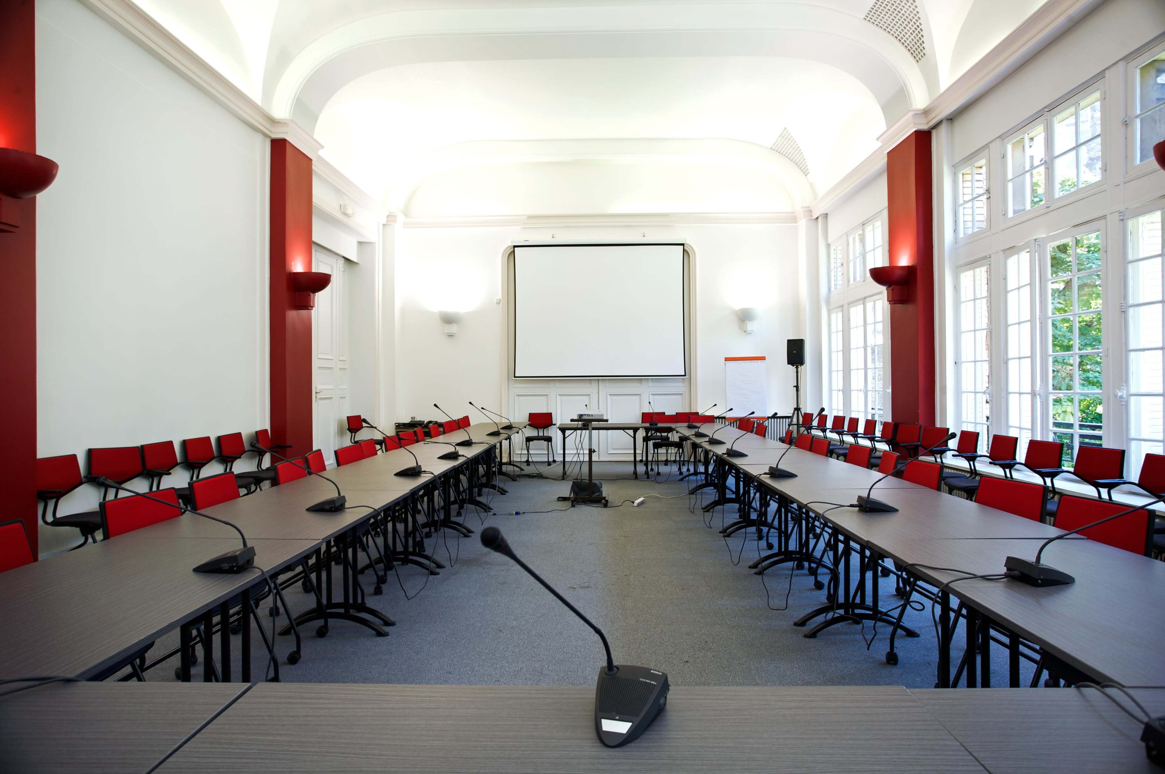 A spacious conference room arranged with multiple tables in a U-shape, surrounded by red chairs, and featuring a large screen at the front.