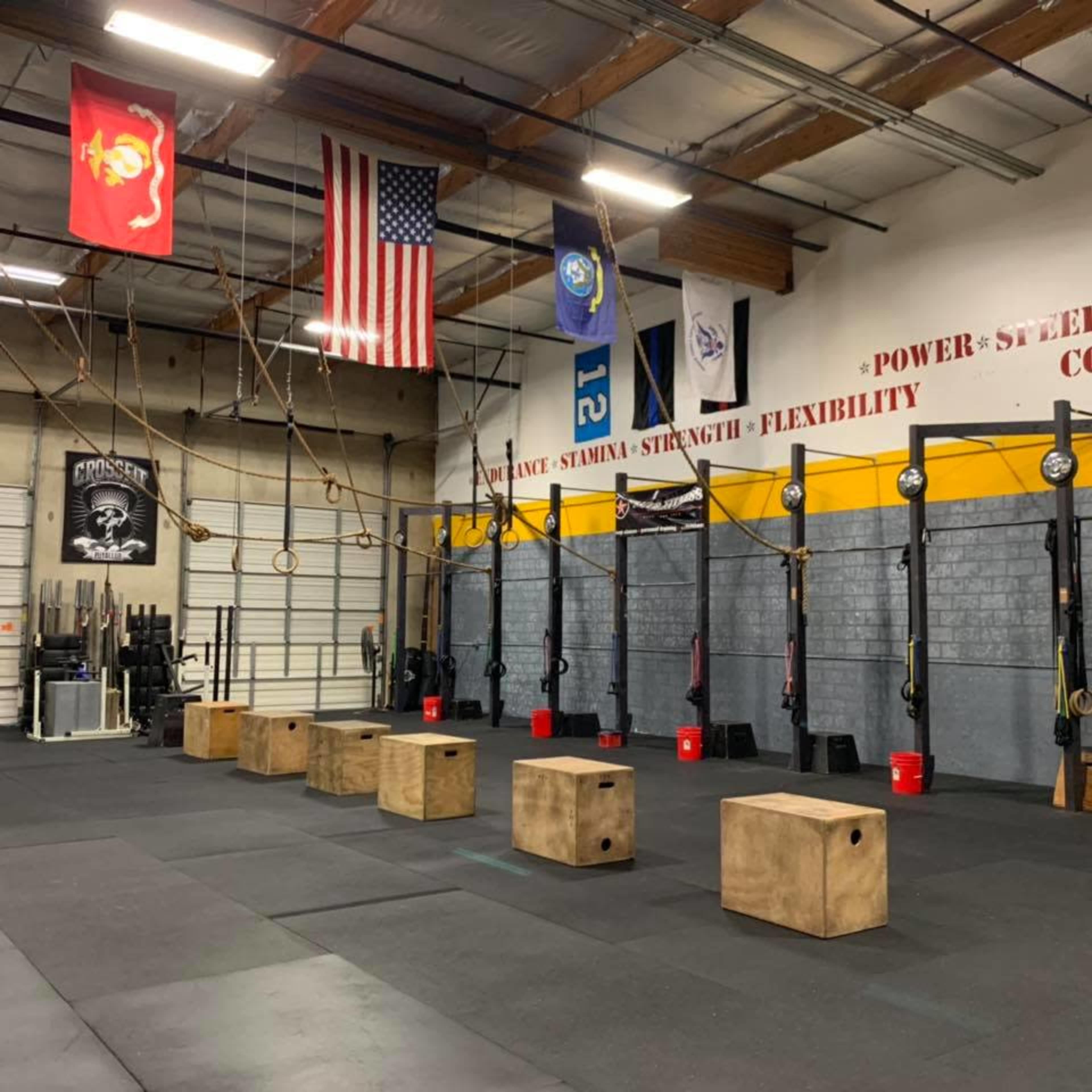The image shows an indoor gym space equipped with several exercise boxes, hanging gymnastic rings, and various flags displayed on the walls.